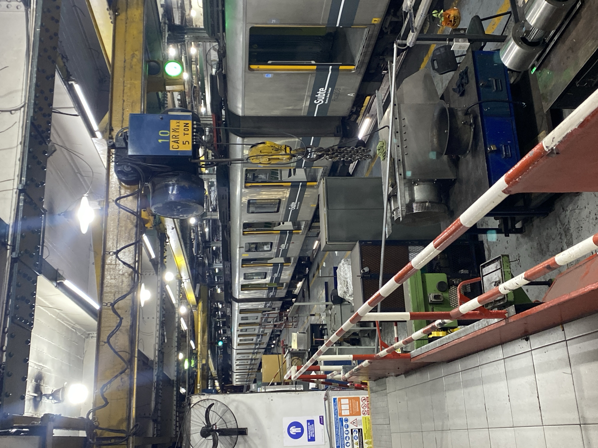 The image depicts the interior of a maintenance facility for the Buenos Aires Metro (Subte). A train car, painted gray with yellow stripes and the "Subte" logo, is visible. A crane with a blue motor block labeled "CAR MAX 5 TON" hangs above the train. The facility features steel beams and structural elements, exposed electrical wiring, and fluorescent lighting. A barrier with red and white stripes runs along a raised platform. A fan is mounted on the wall. Safety signage is posted near the platform, indicating required PPE. The environment is industrial, with evidence of routine maintenance activities. The location can be inferred to be within the city of Buenos Aires, Argentina.
