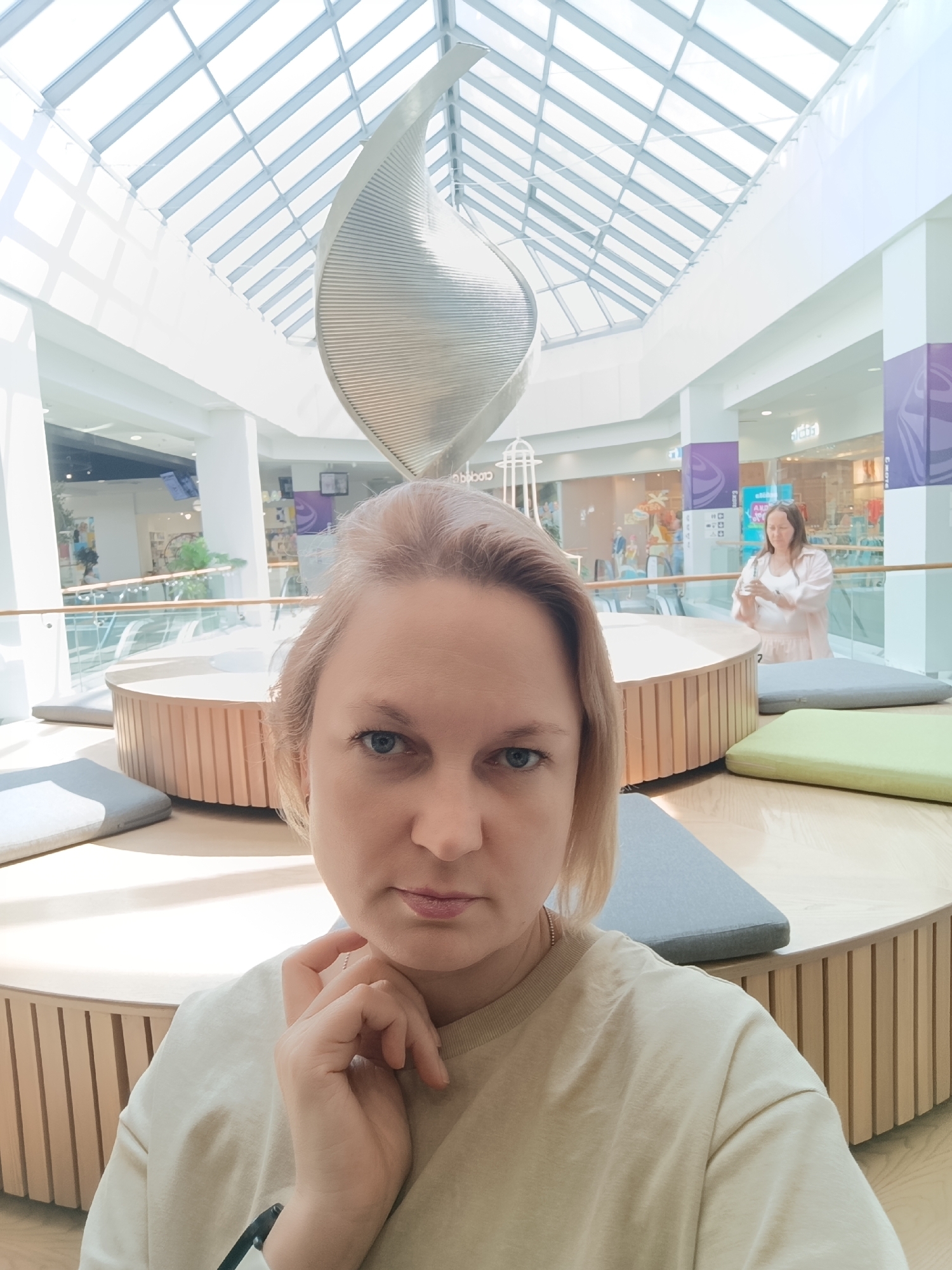 The image shows a fair-skinned woman with short, light-colored hair, blue eyes, and a serious expression. She is positioned in the foreground, with her hand near her chin. She is wearing a light-colored t-shirt. Behind her is a brightly lit indoor space with a high, geometrically patterned skylight. A modern, abstract sculpture sits centrally in the background. A circular wooden structure with cushions arranged around it is visible. Another woman is standing in the background, near a glass railing. The location is the shopping mall "Taltsman" in Izhevsk, Russia.