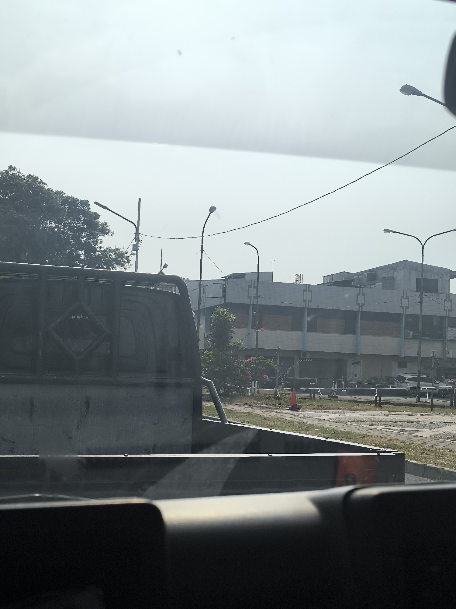 The image captures a view from inside a vehicle, looking out towards a street scene in Tangerang, Indonesia. In the foreground, the interior of the vehicle, specifically the back of a truck bed, is visible. The truck bed appears to be made of dark metal and has some wear and tear.
Beyond the truck, a two-story building with a light-colored facade dominates the midground. The building has a repeating pattern of dark rectangles on its upper section and appears to have some windows boarded up with wooden planks. There are several streetlights lining the road in front of the building.
To the left of the building, a large tree with dense foliage is present. In front of the building, there is a paved area with some bollards and white tape, possibly indicating a construction or maintenance zone. A bright orange traffic cone is also visible.
Further down the street, a white SUV is parked. The sky is overcast and hazy, suggesting a cloudy or polluted day. The overall impression is that of a typical urban environment in a developing city, with elements of infrastructure and construction visible. The specific location within Tangerang is not identifiable from the visual information provided.