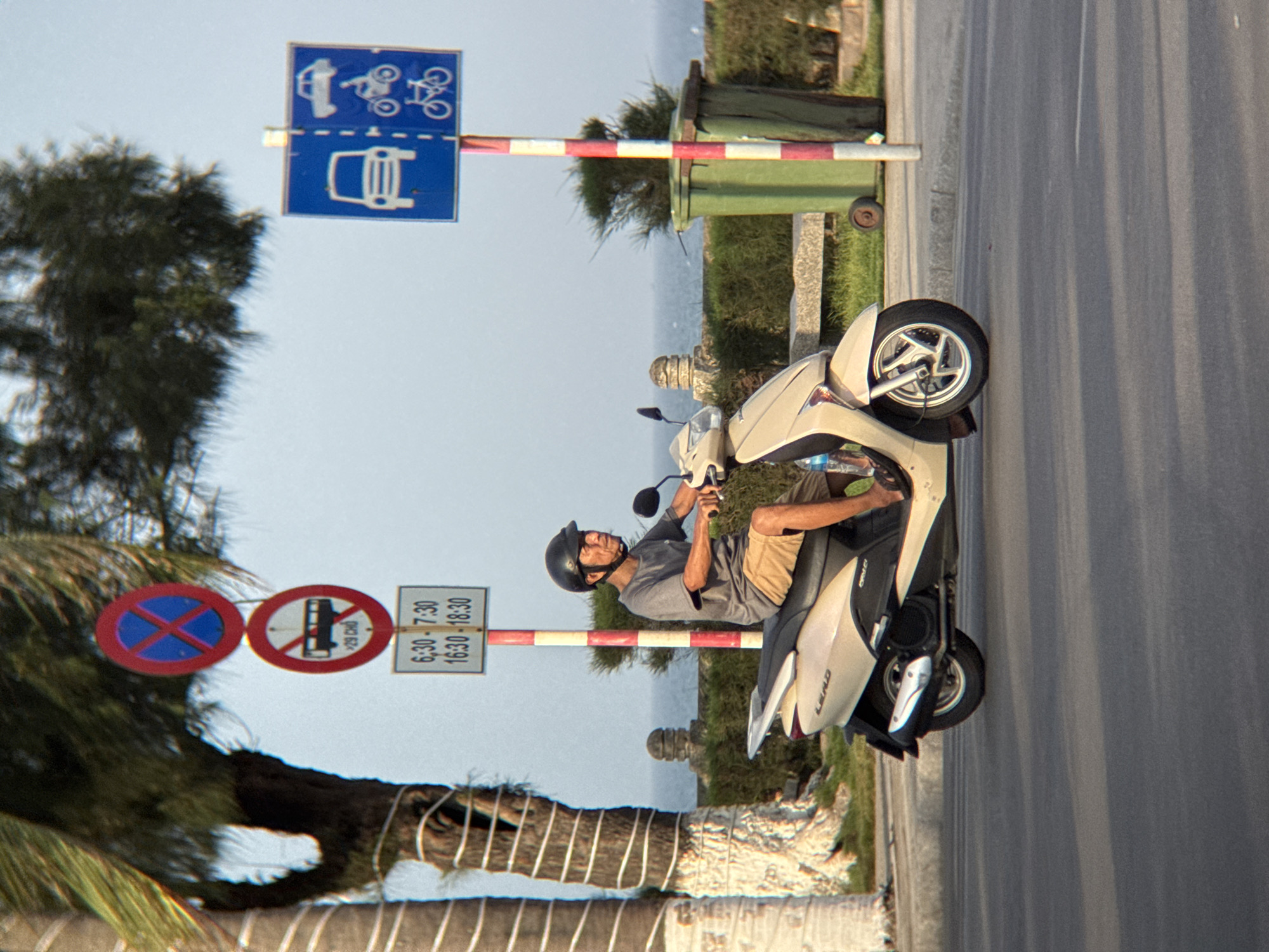 A man wearing a grey t-shirt, khaki shorts, a black helmet, and black sandals rides a beige scooter on a paved road in Nha Trang, Vietnam. The scooter has a black exhaust pipe and silver wheels. The man is looking to his right, with his hands on the handlebars. He is wearing a helmet and has a bottle of water attached to the left side of the scooter. In the background, there are traffic signs, trees, and the ocean. One sign is red and blue with a white X, indicating no stopping. Another sign is red and white with a bus inside a circle and a line through it, indicating no buses. Below this is a sign with times: 6:30-7:30 and 16:30-18:30, likely indicating restricted hours for buses. Another sign to the right is blue with white images of a car and two bicycles, indicating allowed vehicles. A green trash bin with red and white stripes is also visible in the background. The scene appears to be a street in Nha Trang, a coastal city in Vietnam.