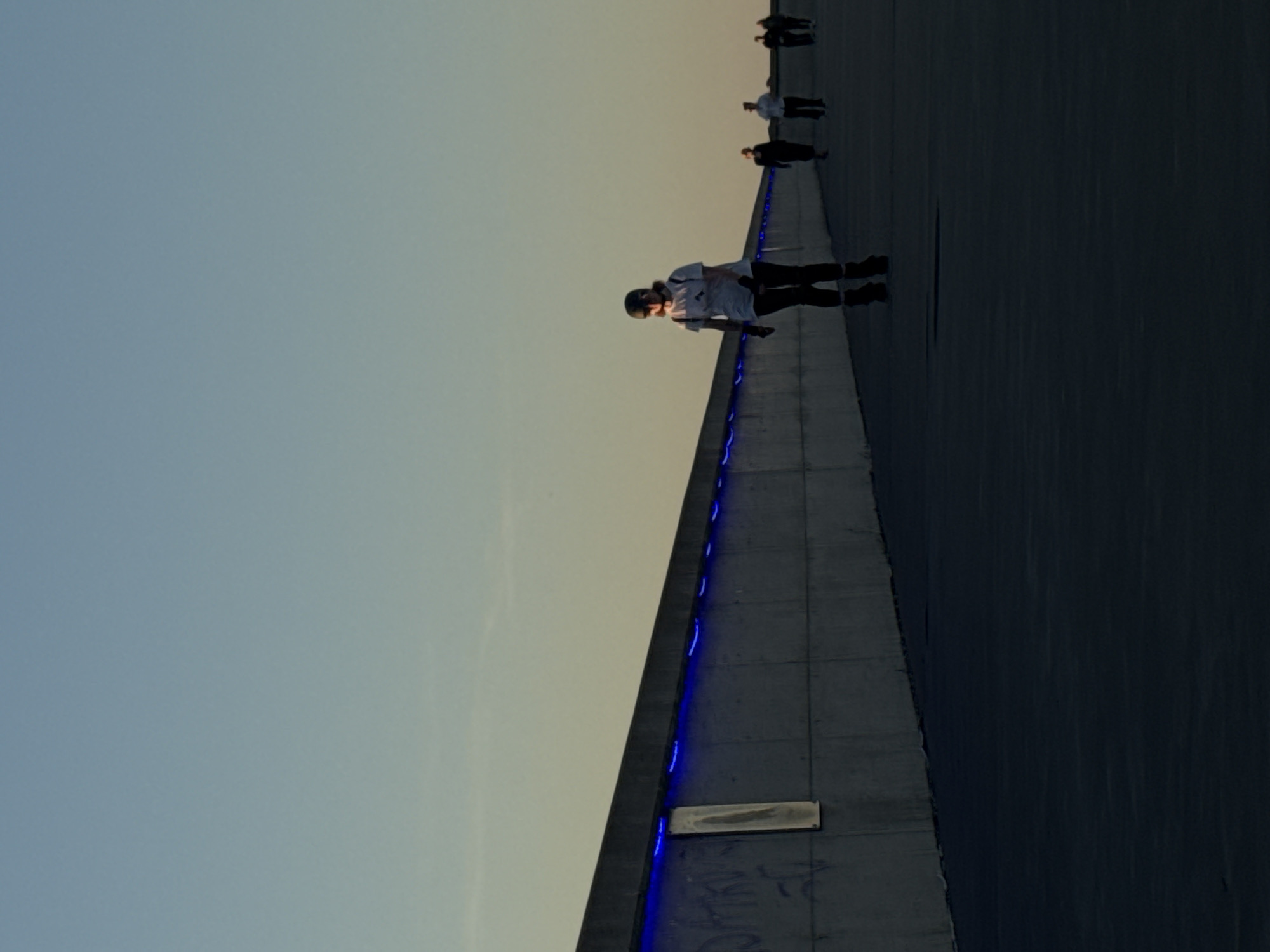 The image depicts an outdoor promenade or walkway under a clear sky displaying a gradient from pale blue-grey at the top to a warm, pale yellow-orange at the horizon on the right, suggesting twilight conditions. A wide, dark paved surface borders a lengthy concrete wall that recedes diagonally from the bottom-left toward the right. This wall is illuminated along its top edge by a continuous strip of blue LED lighting, casting a distinct blue glow. Graffiti in dark lettering is visible on the wall's lower sections, particularly on the left, next to a rectangular light-colored panel.
Multiple individuals are present on the promenade. The most prominent figure, positioned slightly right of center in the mid-ground, is a person wearing a white t-shirt, dark trousers, a dark helmet, and rollerblades. This individual also has a dark backpack and is moving away from the viewer towards the right, with their face illuminated by warm, low-angle sunlight. Further down the path, to the right, are four additional individuals, also moving in the same general direction. One person wears dark attire, followed by another in a light shirt and dark trousers. Two more figures are visible in the distance, partially obscured by shadows. The scene is consistent with the modern waterfront promenade along Reidi tee in Tallinn, Estonia.
