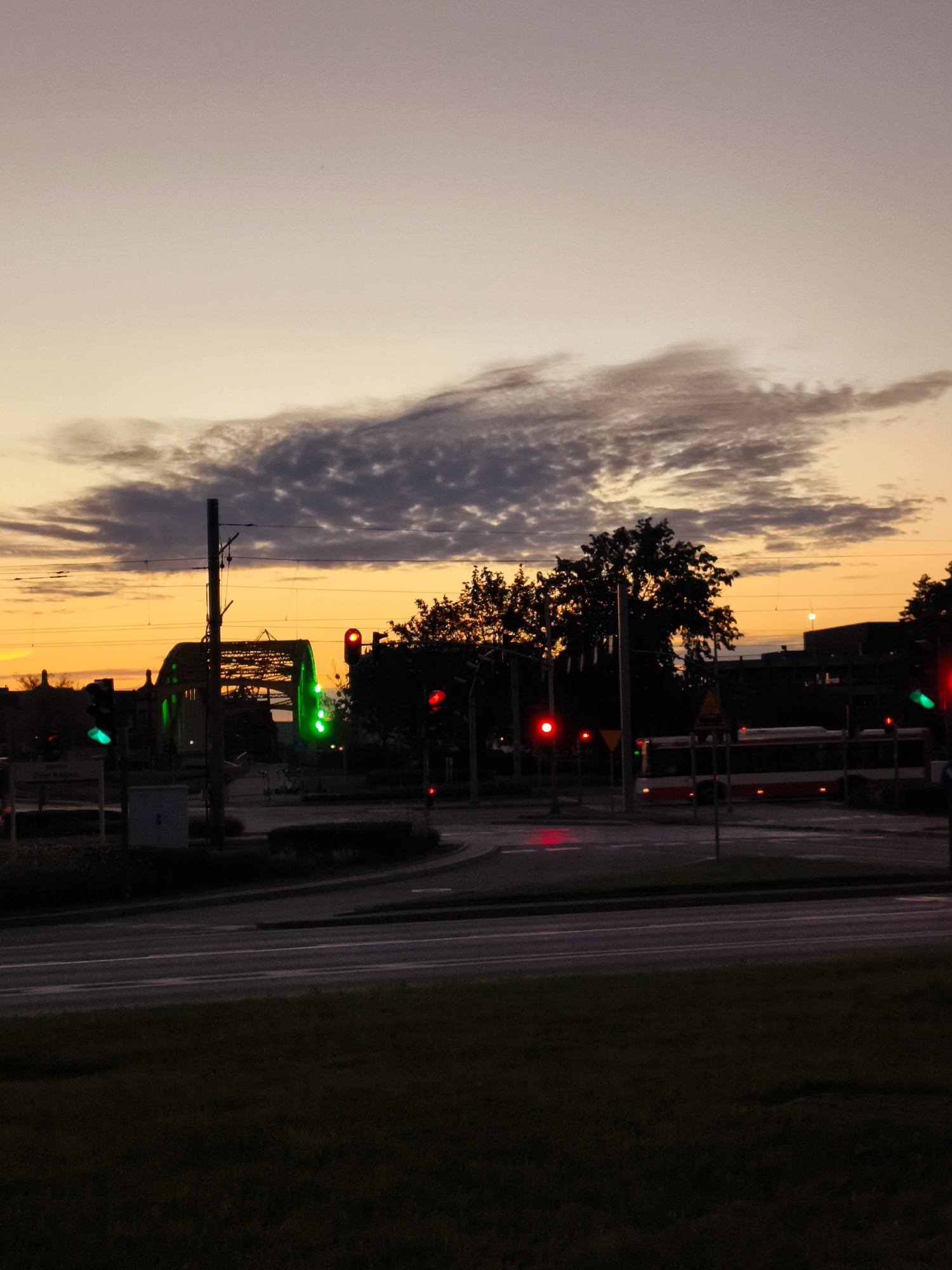 The image depicts an urban scene at dusk, likely in the city of Gdańsk, Poland. The sky transitions from a pale yellow near the horizon to a muted orange and then a soft gray, with scattered dark clouds. A prominent feature is a yellow, arched bridge structure silhouetted against the sky. Traffic lights are visible, with several showing green lights, indicating traffic flow. A bus with a red stripe is driving on the road, also illuminated by traffic signals. Trees line the background, their forms darkened by the fading light. In the foreground, a strip of dark grass is visible. The scene suggests a functioning city street during the evening transition from day to night.