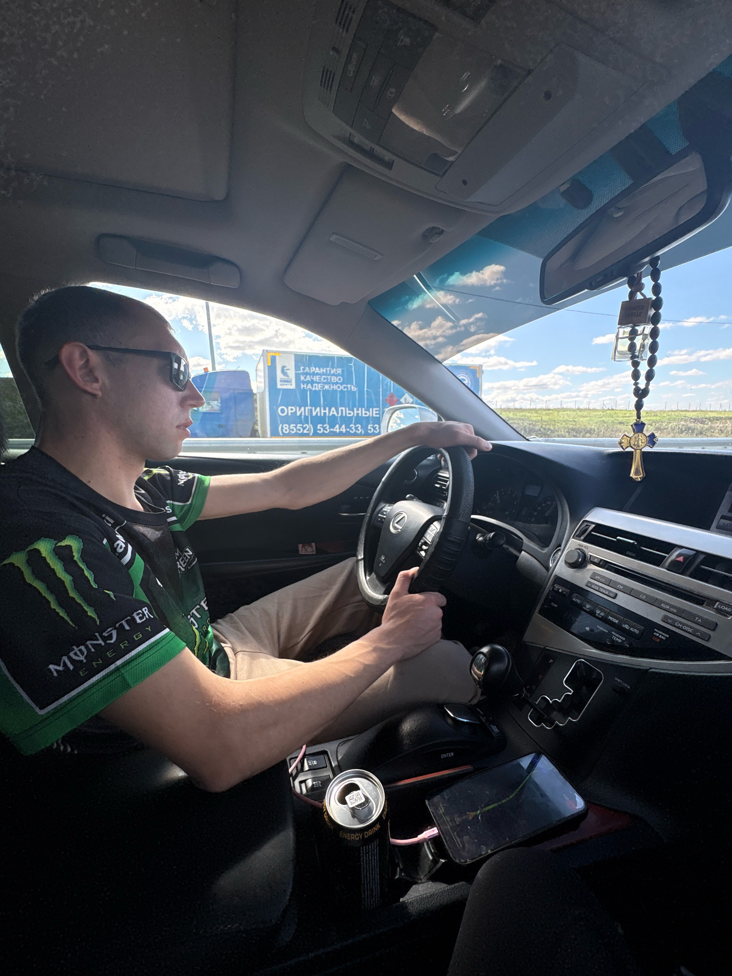 A man is driving a car on a sunny day. The car's interior is visible, including the dashboard, steering wheel, gear shift, and a cell phone. The man is wearing sunglasses and a green and black t-shirt with a Monster Energy logo. There is a can of energy drink in a cup holder. Outside the car, a blue truck with Cyrillic text is visible, along with a field and a bright blue sky with clouds. The scene appears to be on a road near Patrikeevo, Russia.