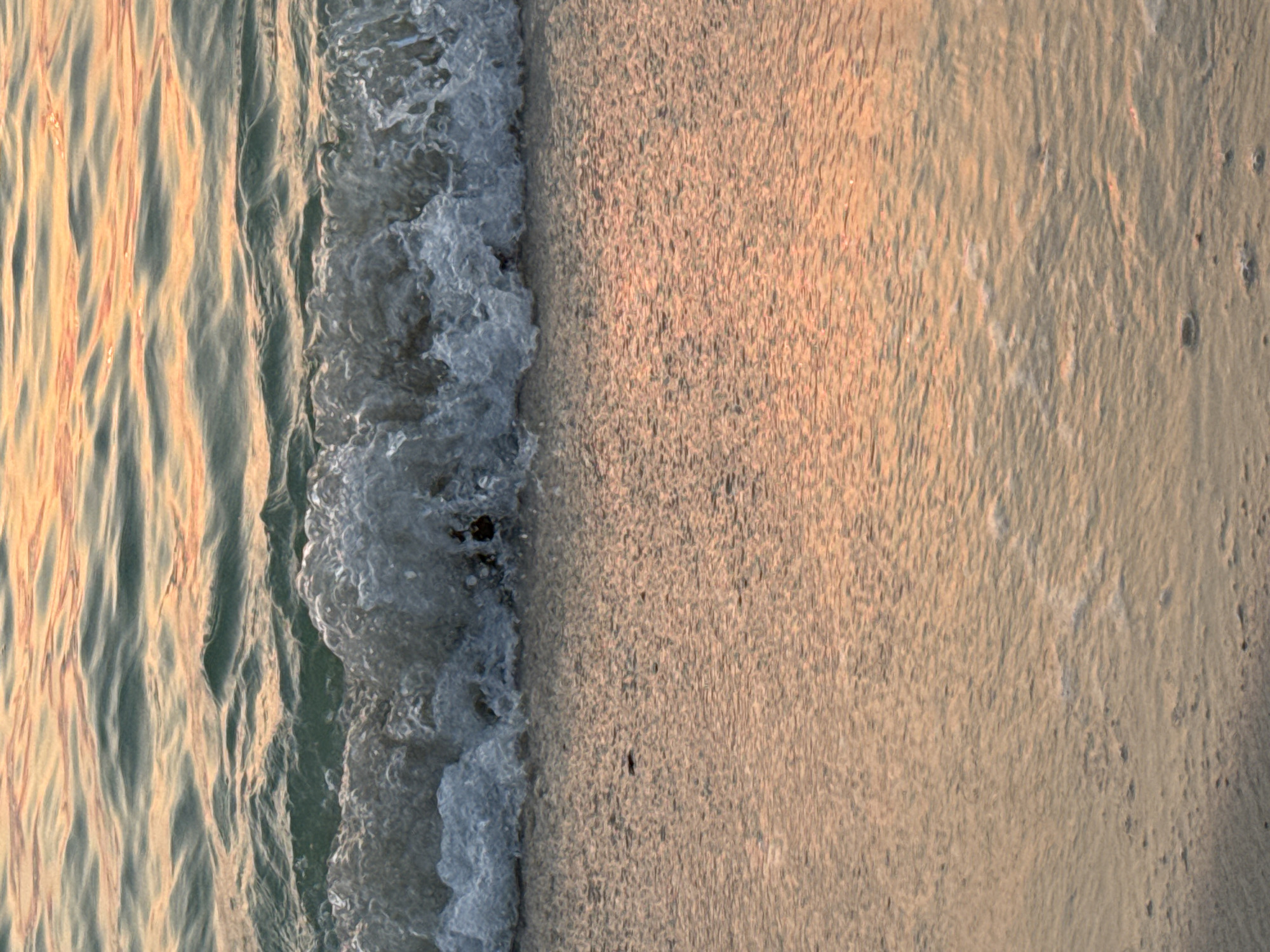 The image captures a close-up view of the sea at Jumeirah Beach in Dubai, United Arab Emirates. The water is shallow, with a small wave breaking near the shore. The sand is wet and reflects the warm, golden light of the sunset. The waves are calm and gentle, with a slight shimmer on their surface. There are no people or objects visible in the frame. The overall mood is serene and peaceful, evoking a sense of tranquility and natural beauty.