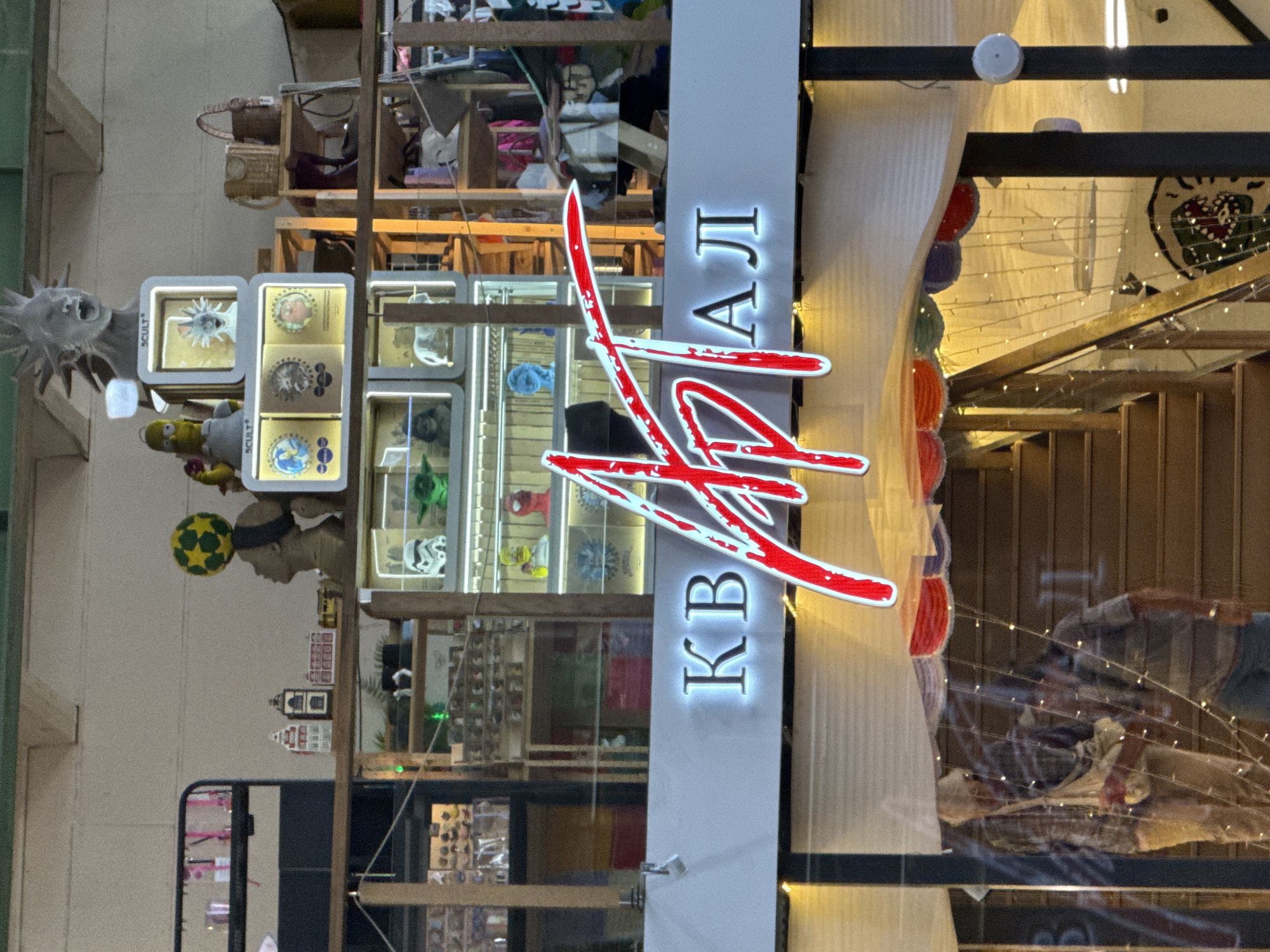 The image shows the exterior of a building with a prominent sign that reads "КВАРТАЛ" in large, illuminated red and white letters. The building appears to be a shop or a cultural space, with various items displayed on shelves in the background. These items include decorative figures like a bust with a spiky hairstyle, Simpsons characters, Star Wars figures, and other assorted curios. There are also baskets, clothing items, and what appear to be miniature houses on display. Two people are visible through a glass partition, one in the foreground holding a bag and wearing a patterned shirt and light-colored pants, and another person behind them. The overall environment suggests a lively and eclectic retail or exhibition space. The sign and the architectural style are consistent with locations in the city of Saint Petersburg, Russia.