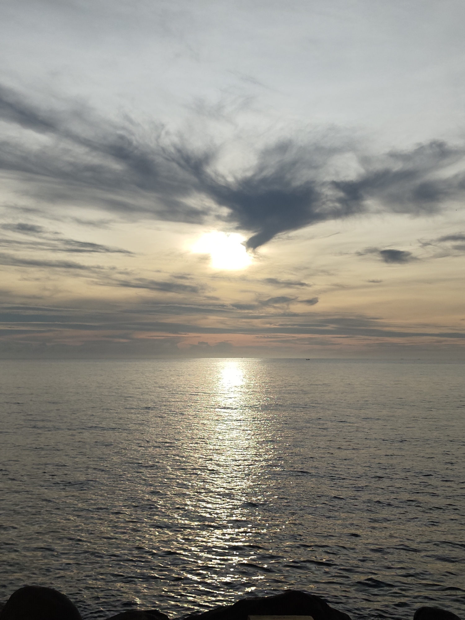 The image depicts a seascape at dusk in Lhokseumawe, Indonesia. The sky is filled with streaky, grey-blue clouds with a large, dark cloud directly obscuring the sun, which shines brightly despite the cloud cover. The sun's light reflects as a shimmering path on the surface of the water. The water is calm with gentle ripples. In the distance, the horizon is visible. The foreground shows a portion of dark, possibly stone, objects that could be part of a breakwater or coastal structure.