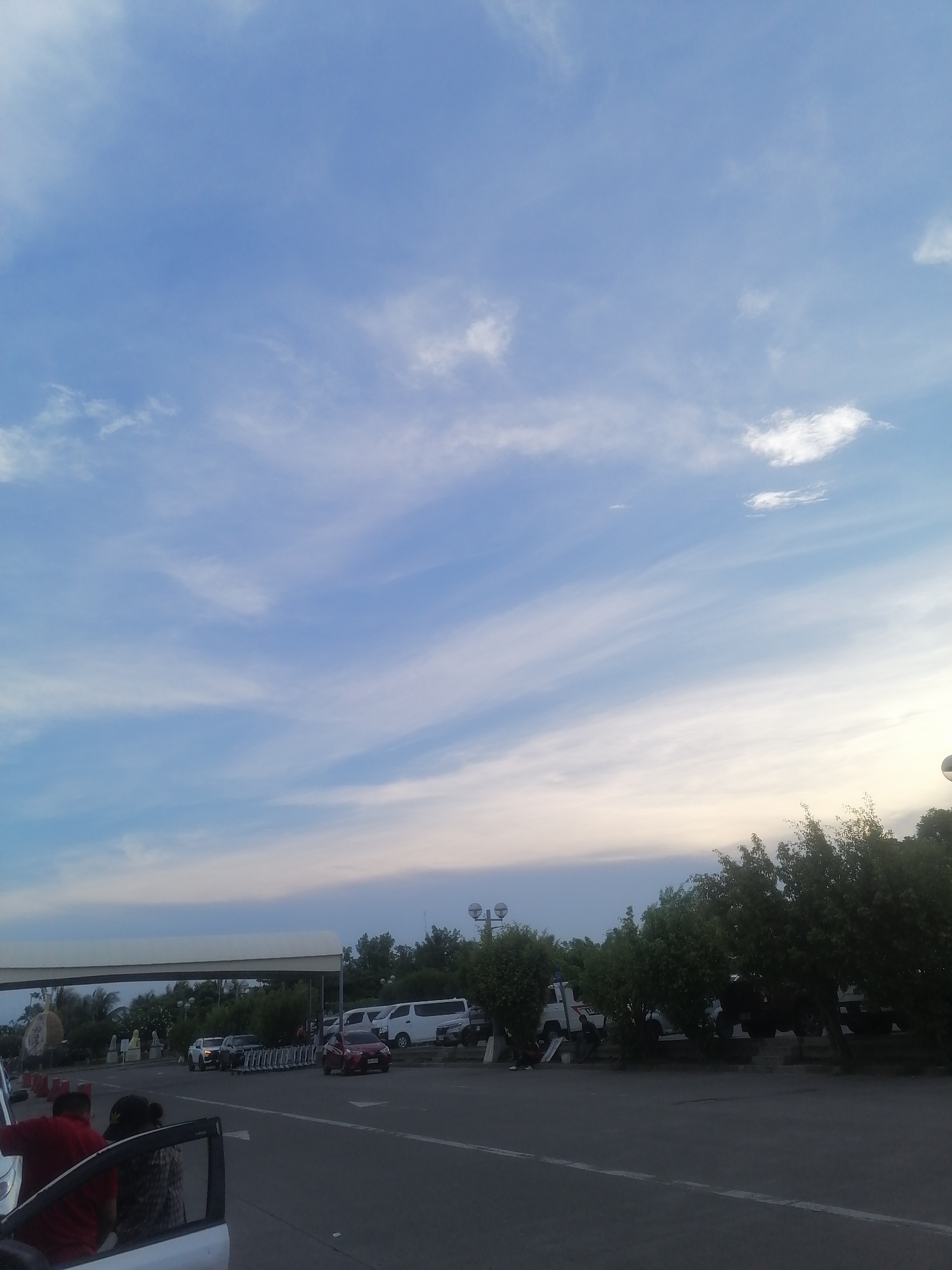 The image presents an outdoor scene likely captured during the twilight hours, either dusk or dawn, in Davao City, Philippines. The expansive sky dominates the upper two-thirds of the frame, displaying a gradient of light blue overhead transitioning to paler, almost white and subtle pastel hues near the horizon, indicative of the changing light. Numerous wispy, elongated white clouds, resembling cirrus or altocumulus formations, are scattered across the blue expanse.
In the foreground, a dark asphalt road with a faded white lane marker stretches across the bottom. On the left side, the open door of a light-colored vehicle, possibly a car or SUV, is partially visible. Two individuals are seen near this open door: one person wearing a red shirt is to the left, and another person with dark hair and a patterned top is partially obscured within or very close to the vehicle's interior.
The midground features a long, white, canopy-like structure or covered walkway extending from the left. Beyond this, a parking area or side road is visible, accommodating several parked vehicles including white vans, a red car, and other light-colored automobiles. Rows of dense green trees and bushes line the area behind the parked vehicles and continue towards the right, suggesting a landscaped boundary or park-like environment. Unlit streetlights are interspersed among the foliage. The overall lighting is soft and muted, contributing to the tranquil early morning or late evening atmosphere. No discernable text is visible on any objects or signs.