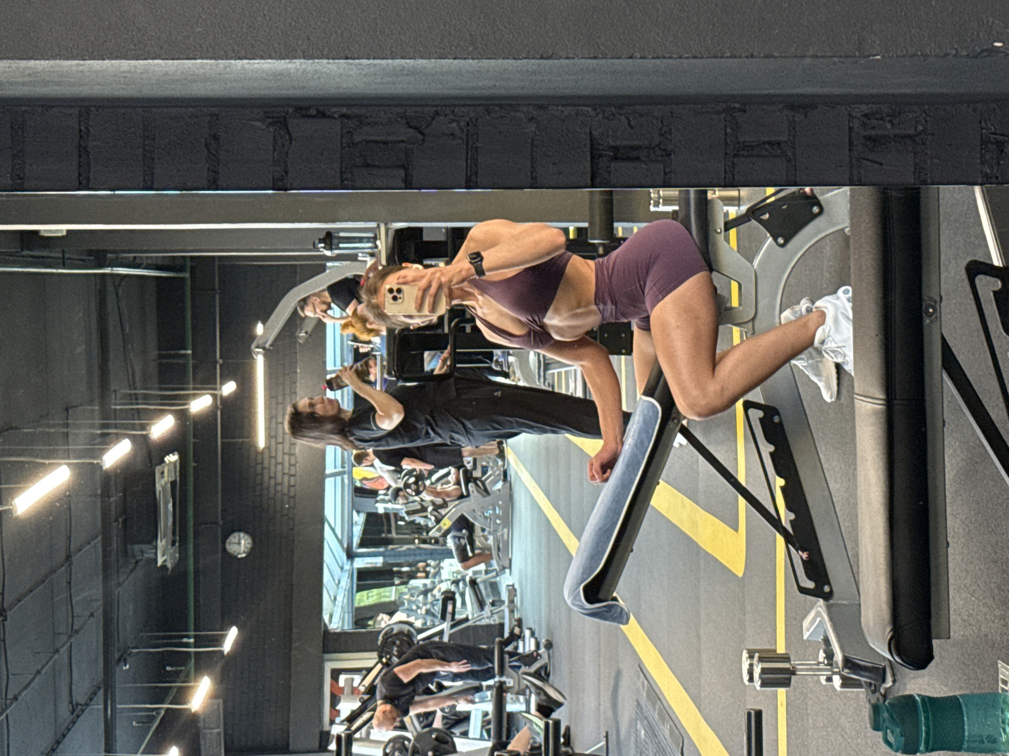A woman in athletic wear is taking a selfie in a gym. She is sitting on a workout bench, facing a mirror. She is wearing a purple sports bra and matching shorts, and white athletic shoes. In the background, other gym-goers are visible, as well as exercise equipment like weight racks and benches. The gym has a concrete floor with yellow lane markings, and industrial-style lighting on the ceiling. The image appears to be taken in Saint Petersburg, Russia, based on similar gym interiors found in the city.