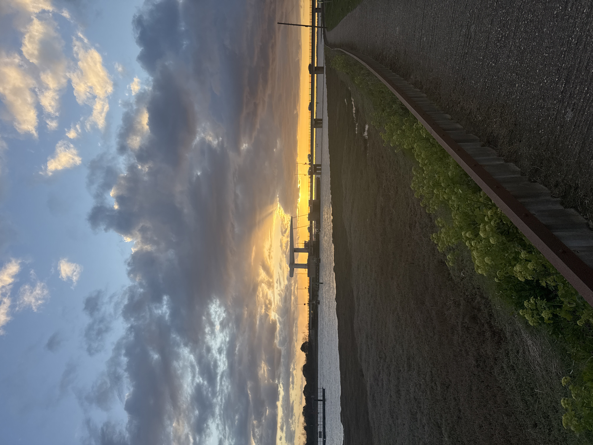 The image shows a sunset over the River Yare in Great Yarmouth, United Kingdom. The sun is setting behind the Haven Bridge, a road bridge over the river. The sky is filled with clouds, some reflecting the golden light of the sunset. To the right of the frame is a path next to the river, with vegetation growing along the edge. The image is taken at ground level, and the camera is angled upwards.