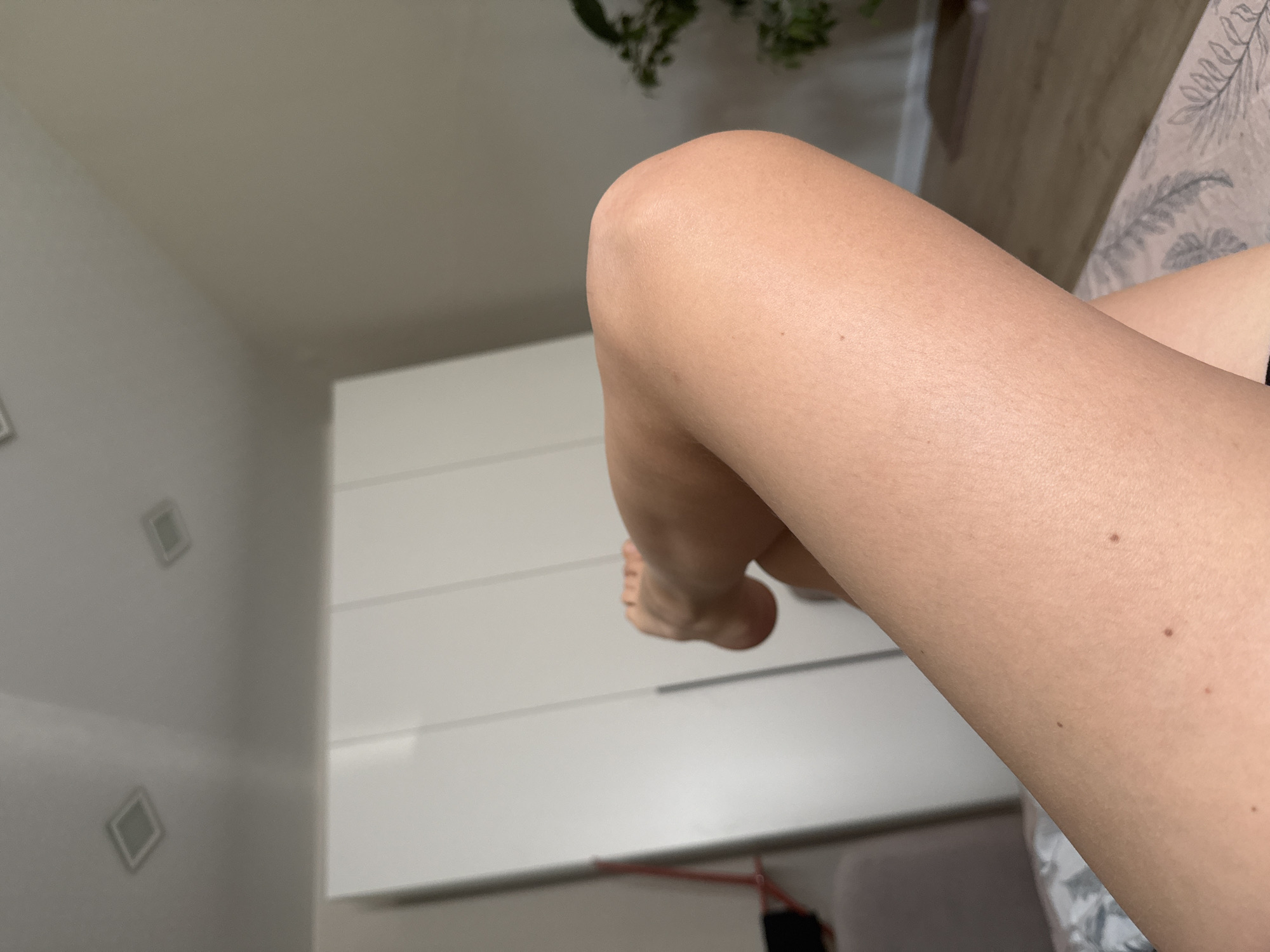 The image shows a person's crossed legs in an indoor setting. The person is lying on a bed with a patterned bedspread. A white dresser is visible in the background, along with a wall with square recessed lighting. A plant is partially visible in the upper portion of the frame. No information indicates the location is in Izhevsk, Russia.