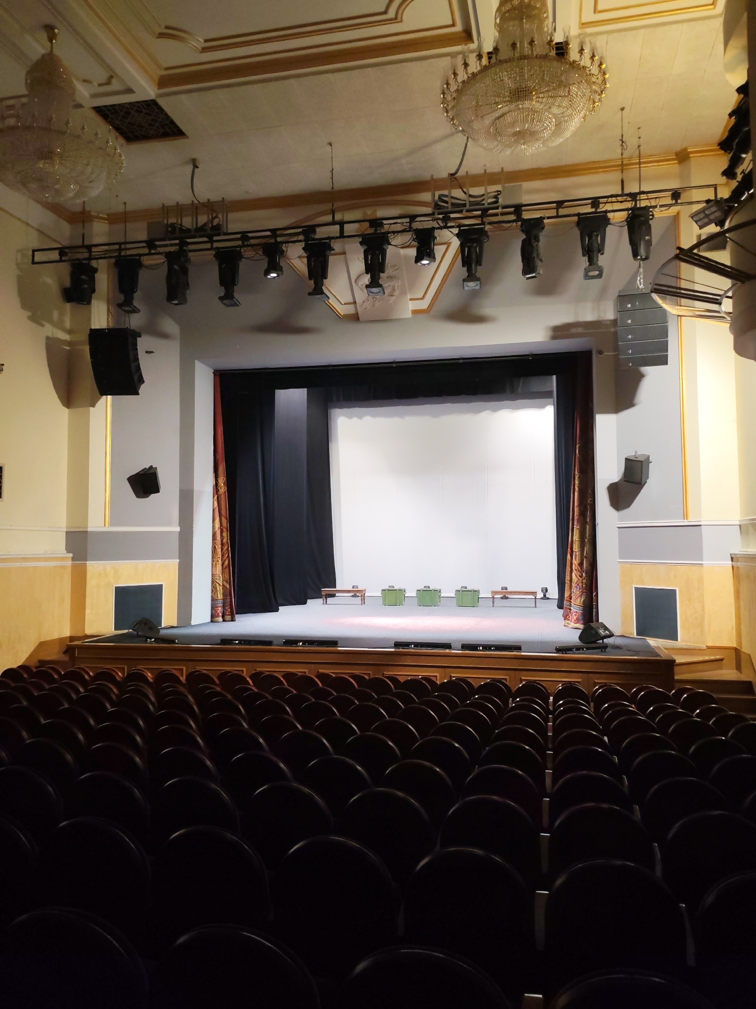The image shows the interior of a theater. Rows of dark seats fill the foreground, suggesting an auditorium setup. The background features a stage with a white screen backdrop and a set of black curtains, flanked by decorative golden and red curtains. Stage lighting equipment is suspended from the ceiling above the stage. Several pieces of furniture are present on the stage, including a small bench and what appear to be green boxes. The architecture of the theater features ornate details, including chandeliers and decorative moldings. Based on this information, this is the State National Theater of the Udmurt Republic located in Izhevsk, Russia.