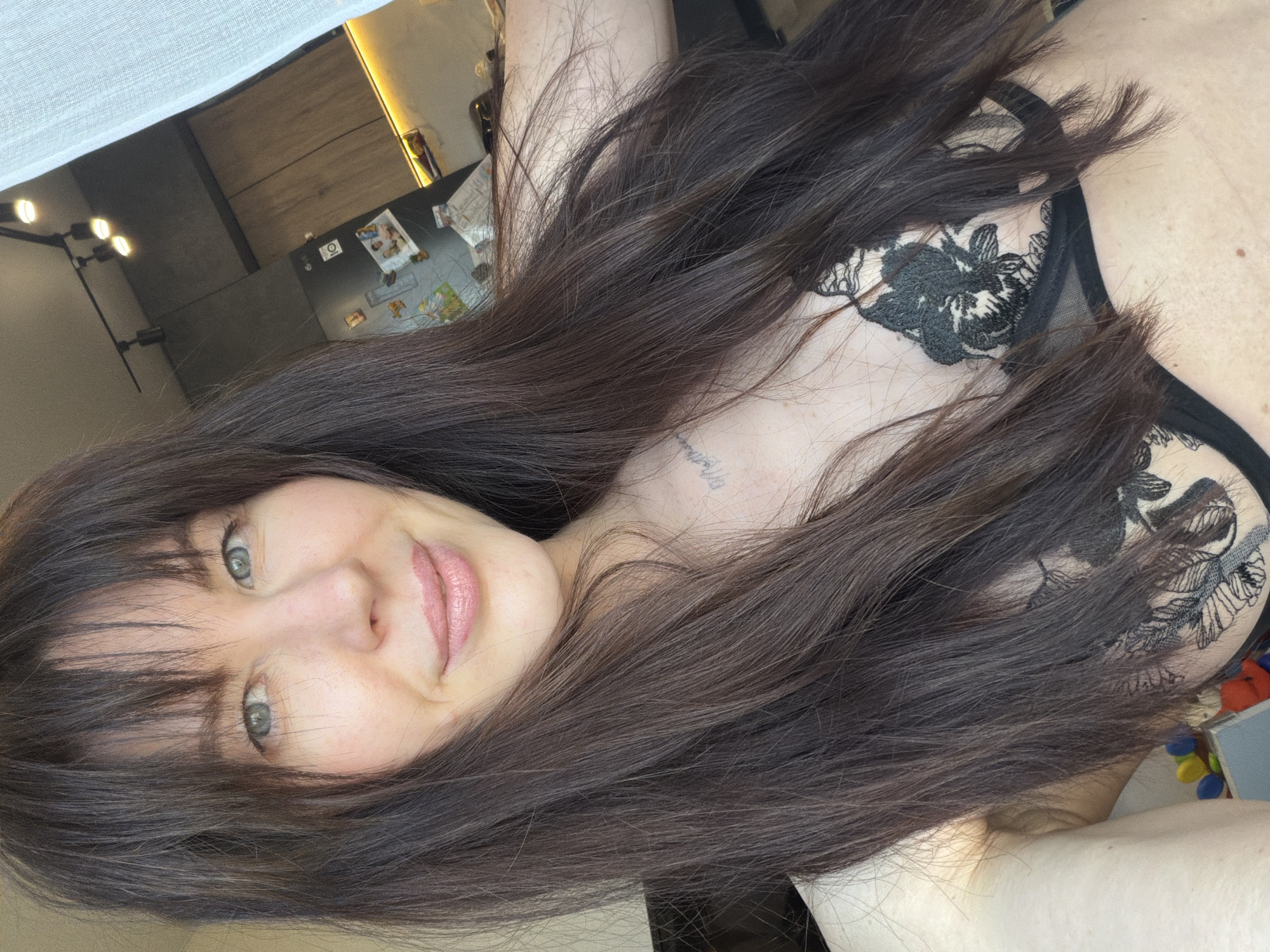 A woman with long, dark hair is shown in a close-up, slightly rotated view. She has fair skin, light-colored eyes, and is wearing lipstick. The image includes a portion of her upper body, revealing a bra with a floral design. A tattoo is visible on her collarbone area. The background shows an interior space, including a kitchen area with cabinets, a refrigerator with items attached, and lighting fixtures. The location is presumed to be Perm, Russia, but there is no specific information to verify the exact city or location.