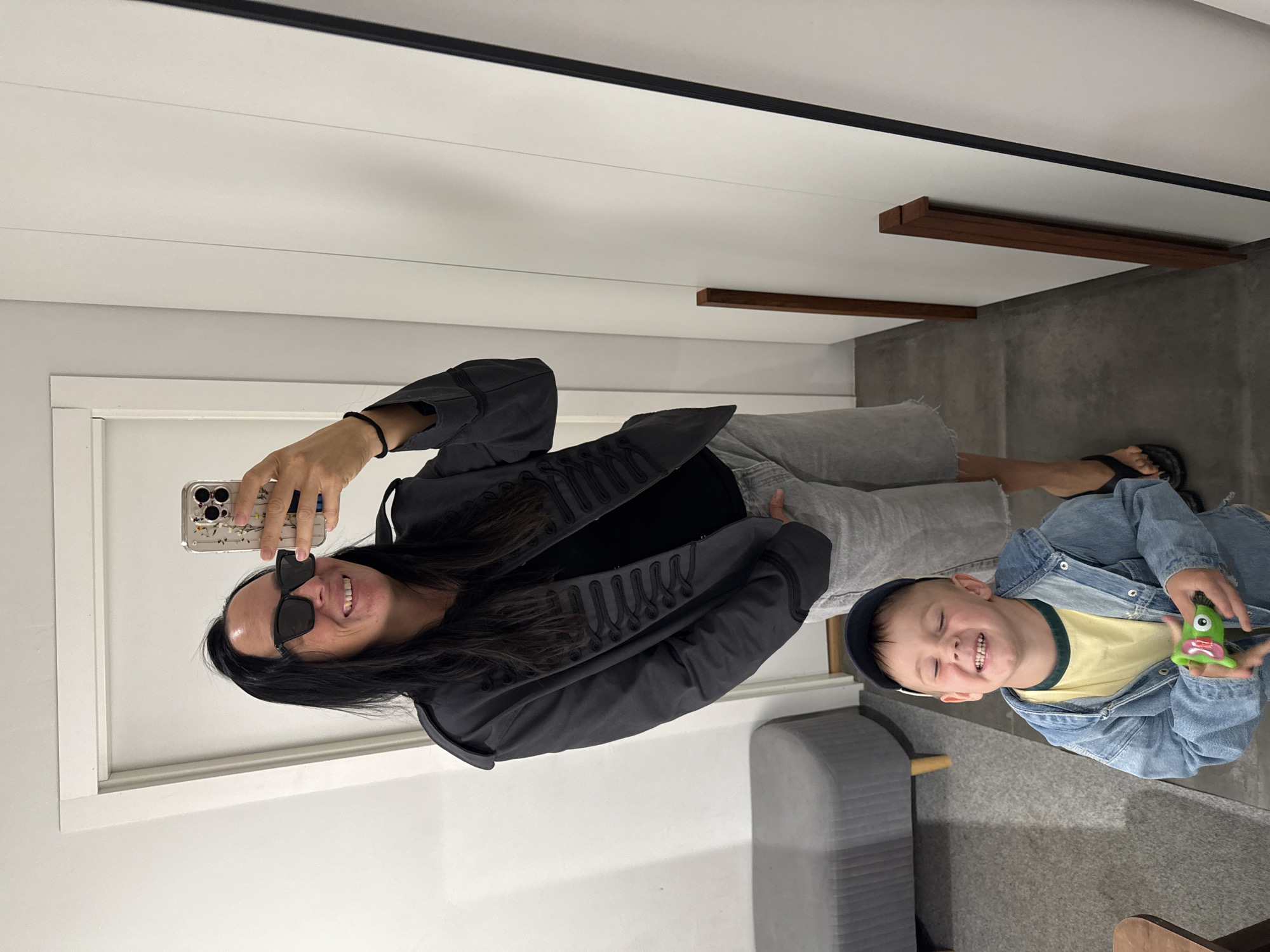 The image shows a woman in a grey military-style jacket, black t-shirt, and grey shorts taking a mirror selfie. She is wearing sunglasses and has long dark hair. A young boy in a denim jacket and a blue baseball cap is in the foreground, smiling and holding a green toy. The scene appears to be an indoor dressing room or fitting area, with a bench, a door, and a large mirror. There is no indication of the location being Chaikovsky, Russia.
