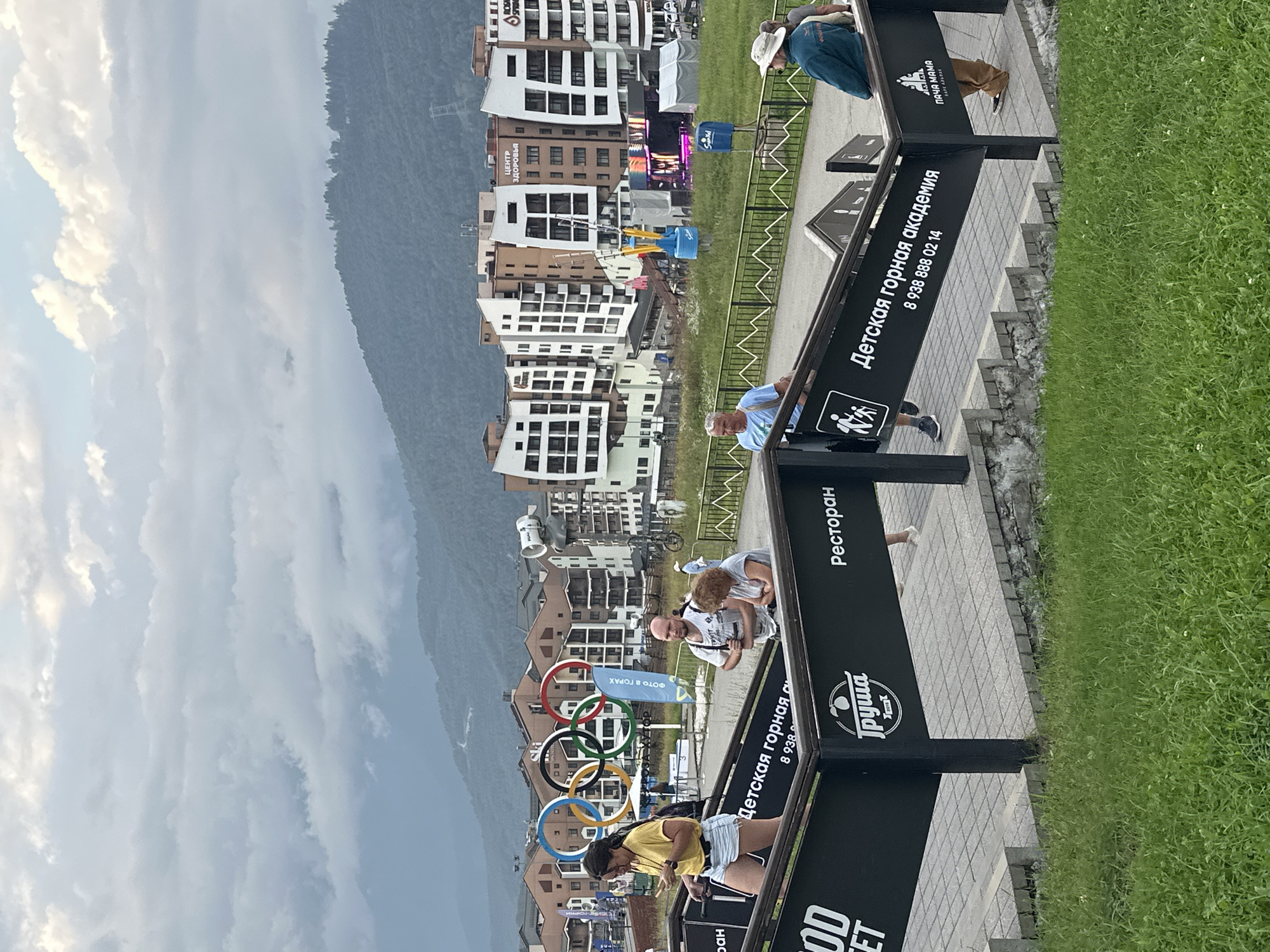 In Estosadok, Russia, this image captures a cloudy day with a backdrop of lush green mountains and modern apartment buildings. The scene is set on a set of outdoor stairs, with several individuals ascending. A woman in a yellow shirt and denim shorts walks up the stairs, looking at her phone. To her right, a bald man with a backpack stands with his hands clasped. Further up, a woman with auburn hair leans on the railing. At the top of the stairs, a man in a blue jacket and a wide-brimmed hat is visible, along with other people in the background. Prominently displayed along the staircase are black signs with white text. One sign reads "Детская горная академия" (Children's Mountain Academy) followed by a phone number. Another sign features a logo of a pear and the word "Груша" (Pear) with "Ресторан" (Restaurant) below it. In the distance, the Olympic Rings are a recognizable landmark, set against the backdrop of the mountainous resort. The architecture of the buildings is modern, with a mix of white and brown facades. Various flags and banners are scattered throughout the scene, indicating a lively atmosphere. A tent structure and a stage with lighting are also visible in the mid-ground, suggesting an event is taking place or has recently concluded. The overall impression is of a bustling mountain resort town, with a blend of natural beauty and man-made attractions.