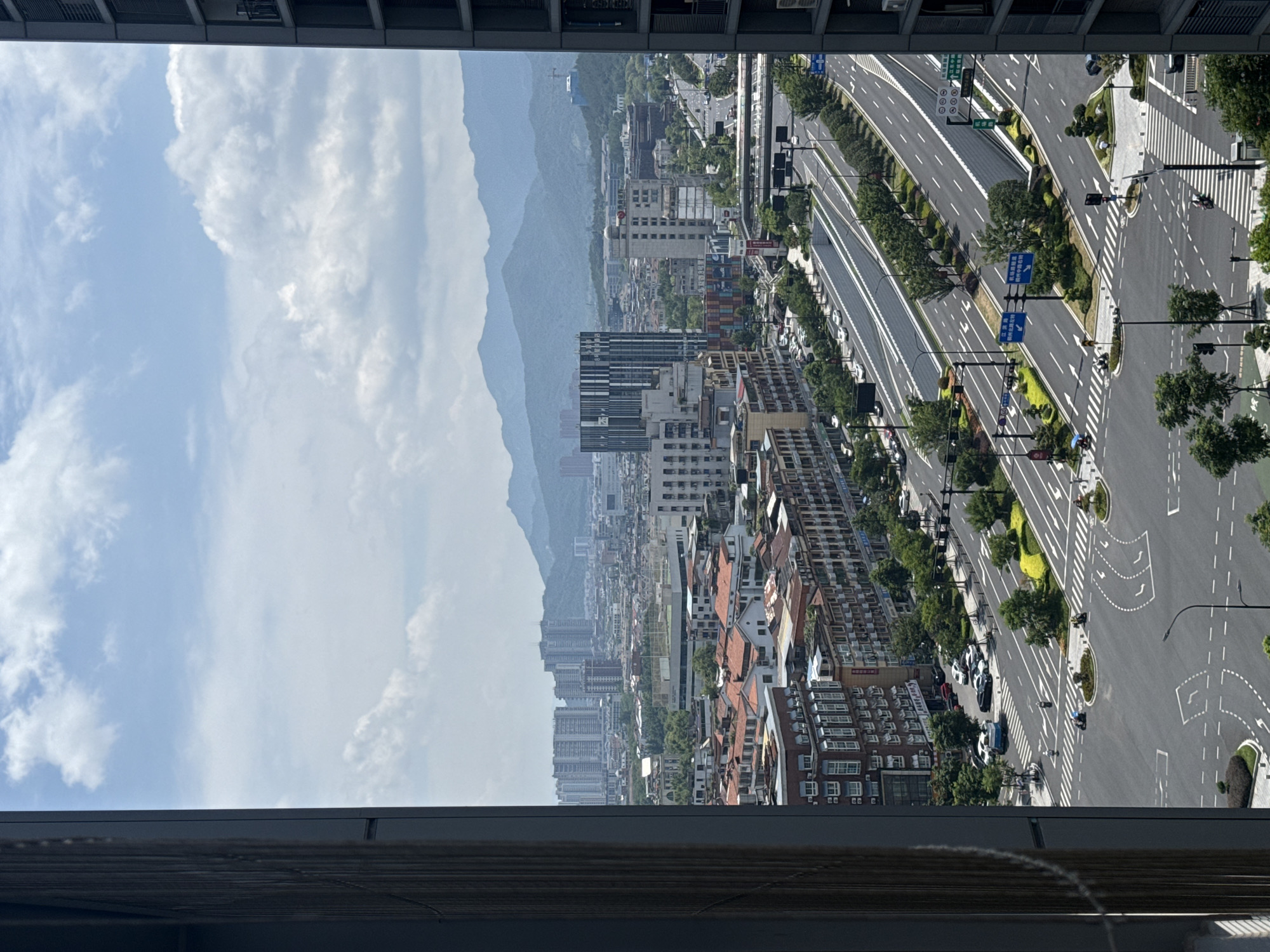 This image captures a panoramic view of Yiwu, China, showcasing a bustling urban landscape dominated by modern architecture and extensive infrastructure. The foreground is framed by the dark, vertical elements of a building on the right and a similar structure on the left, hinting at the perspective from a high vantage point, possibly an apartment or office window.
The scene unfolds across a city with a distinct blend of residential and commercial buildings, many featuring red-tiled roofs that add a traditional touch amidst the contemporary structures. In the mid-ground, a multi-lane highway system is prominently displayed, featuring well-maintained asphalt surfaces, clear lane markings, and traffic signs. The highway appears to be elevated in some sections, with ramps and underpasses indicating a sophisticated transportation network designed for efficient movement of vehicles. Trees and green spaces are strategically planted along the roadways and in the medians, adding a touch of nature to the urban environment.
Various vehicles, including cars and scooters, are visible on the roads, suggesting active traffic flow. Pedestrian crossings and traffic lights are also present, indicating a well-regulated urban setting. Several buildings stand out, including a tall, modern skyscraper with a dark, striped facade in the mid-ground, and other office buildings and residential complexes of varying heights and architectural styles scattered throughout the cityscape.
In the background, a range of hazy blue mountains forms a scenic backdrop to the city, suggesting Yiwu's location within a geographically diverse region. The sky is a mix of blue with scattered white clouds, indicating a partly cloudy day. The overall impression is one of a dynamic and developing city, characterized by its extensive road network, diverse architecture, and proximity to natural landscapes.