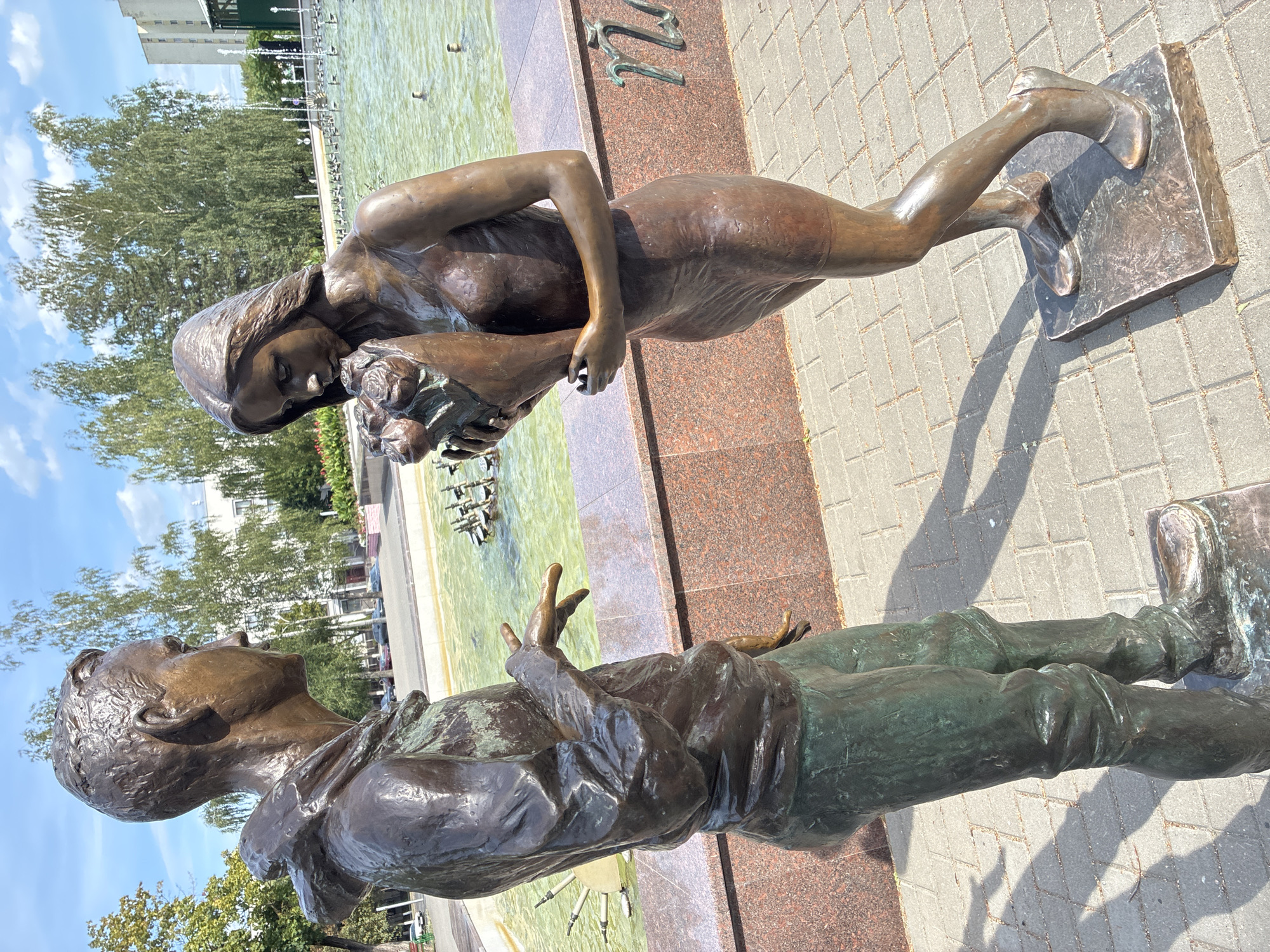 The image shows a bronze sculpture of a young man and a young woman standing in front of a fountain in Mahilyow, Belarus. The young man, on the left, is wearing a hoodie and pants and is looking at the young woman. He has his right hand outstretched, with his fingers slightly splayed, as if speaking or gesturing. The young woman, on the right, is wearing a dress and is holding a bouquet of roses to her face, smelling them. She is looking down at the flowers. The fountain behind them has green water and several spouts of water erupting. The ground is paved with gray interlocking bricks. In the background, there are trees, buildings, and a blue sky with some clouds. The overall scene depicts a romantic moment between two young people.