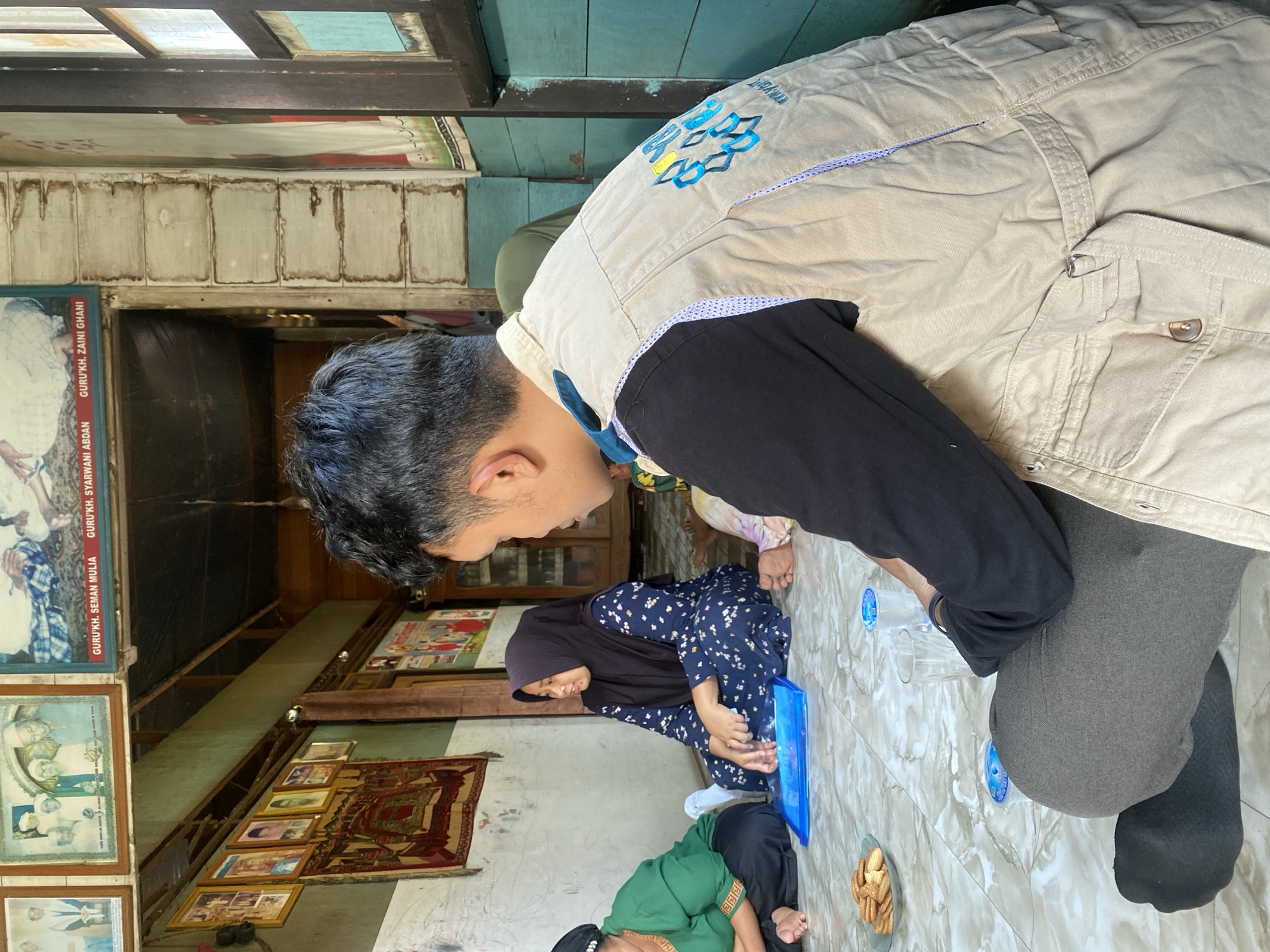 In Bawahan Selan, Indonesia, a man wearing a tan vest with blue and yellow embroidered lettering is seated on the floor, facing away from the camera and looking towards a group of people. The group includes two women sitting on the floor, one in a green top and black pants, the other in a navy blue hijab and floral dress. A small plate of cookies, a glass of water, and a blue tray are on the floor near them. The room appears to be an indoor space with rustic wooden walls and ceiling. Several framed pictures and tapestries hang on the walls, some depicting individuals and group portraits. The overall atmosphere suggests a casual gathering or meeting.