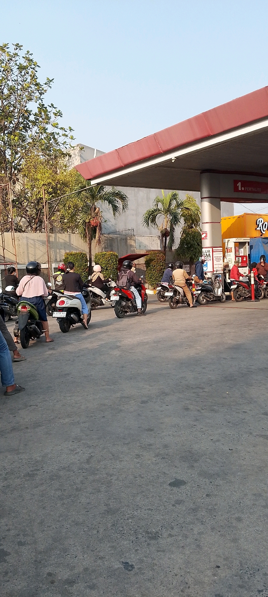 The image depicts a daytime scene at a Pertamina gas station in Bekasi, Indonesia, with a clear sky and early morning or late afternoon sun casting long shadows. Several individuals on scooters are waiting in line to refuel.
In the foreground, the asphalt ground is visible, with a few stray leaves. To the left, a person in blue jeans and grey slip-on shoes stands next to a dark-colored scooter. The scooter's license plate, "B 5138 KGL," is partially visible.
Further back, a line of scooters and riders extends towards the gas pumps. One rider in a pink helmet and light pink top is on a white scooter. Another rider in a black helmet and dark clothing is on a red scooter. A person in a brown uniform is on a black scooter.
The gas station canopy, with a red roof and white trim, covers the pumps. A yellow building with "RO" visible on it is situated behind the pumps, suggesting it is a convenience store or part of the gas station facilities. A sign on a pump column reads "1. PERTALITE." Tropical palm trees and dense green bushes are present in the background, along with a grey concrete wall. The overall atmosphere is that of a busy, routine day at a local gas station.