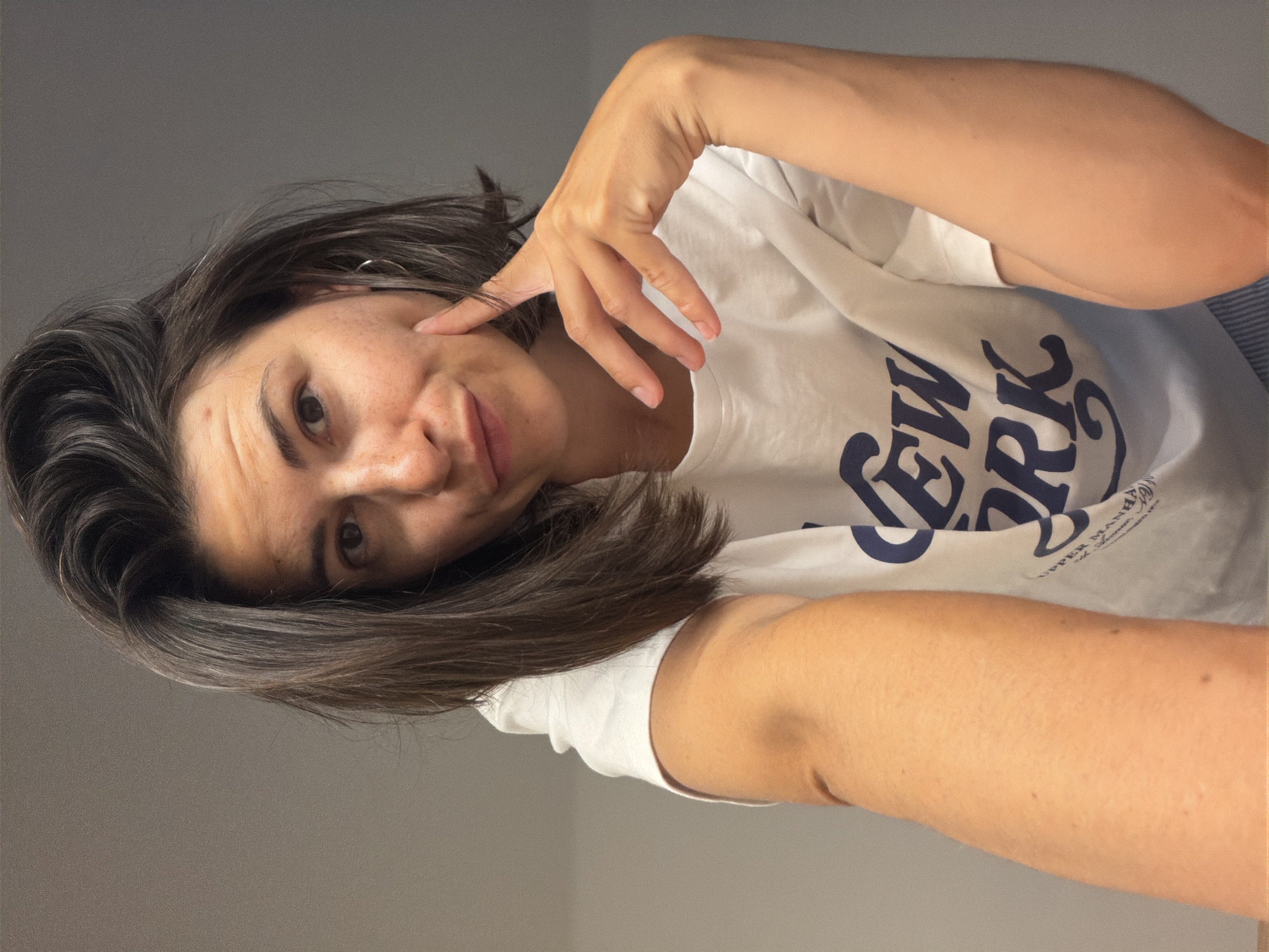A woman is seen in a selfie-style, tilted portrait shot. She has dark hair, fair skin with freckles on her face, and is wearing a white t-shirt with the words "New York" printed on it. Her hand is near her face. The background is a plain gray wall. The location in the city of Budva, Montenegro is not discernible.