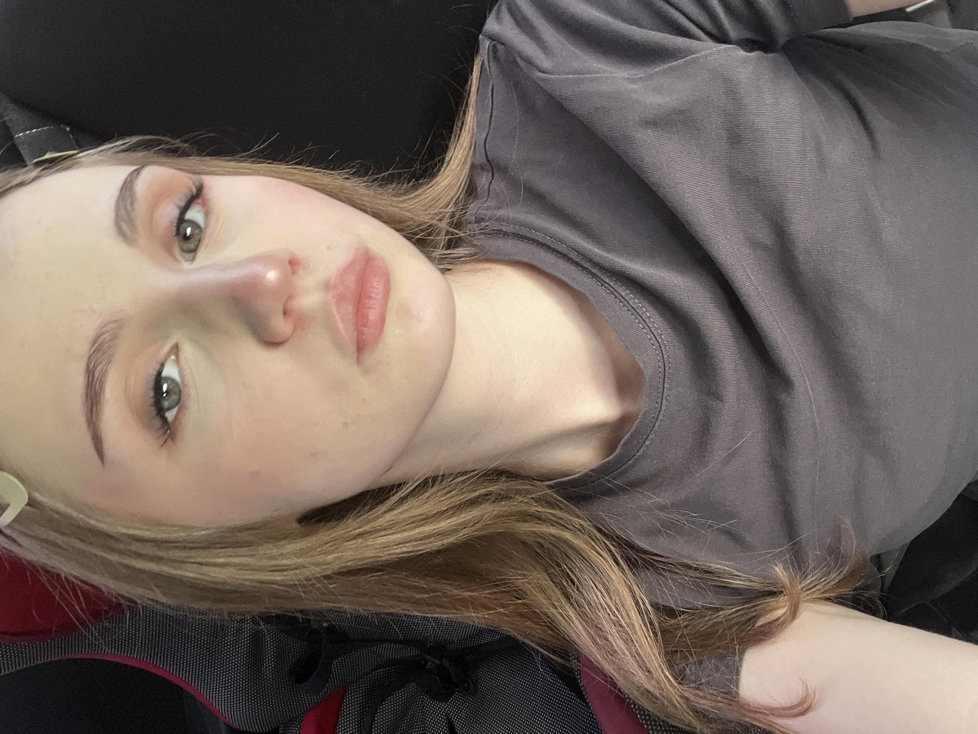 A young woman with blonde hair is captured in a close-up, head-and-shoulders shot. She is looking directly at the camera with a neutral expression. Her eyes are a hazel green, and she is wearing subtle eye makeup. Her lips are slightly parted, and her skin appears smooth with a touch of blush on her cheeks. She has a light-colored hair clip visible in her hair. She is wearing a plain, dark gray t-shirt. The background is a mix of a dark, possibly upholstered surface and a black background, suggesting she might be seated in a chair or in a vehicle. The overall lighting is soft, highlighting her facial features. The image does not contain any information that would allow for its location to be determined within the city of Новая Отрадовка, Russia.