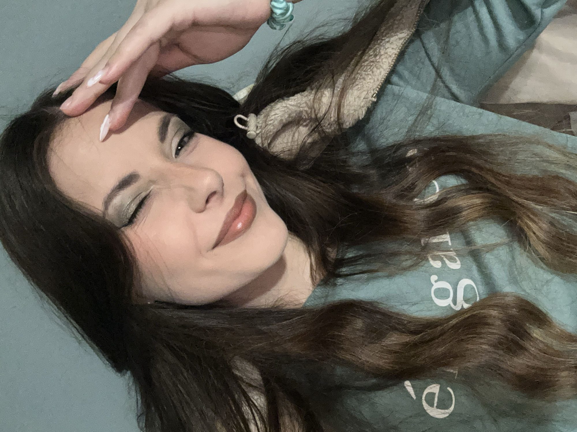 A young woman with long, brown hair is lying down, partially facing the camera, and winking. She is wearing a light green top with white lettering on it and a jacket with a furry lining. Her right hand is resting on her forehead. The background appears to be a plain wall. The location is Unknown, Unknown.