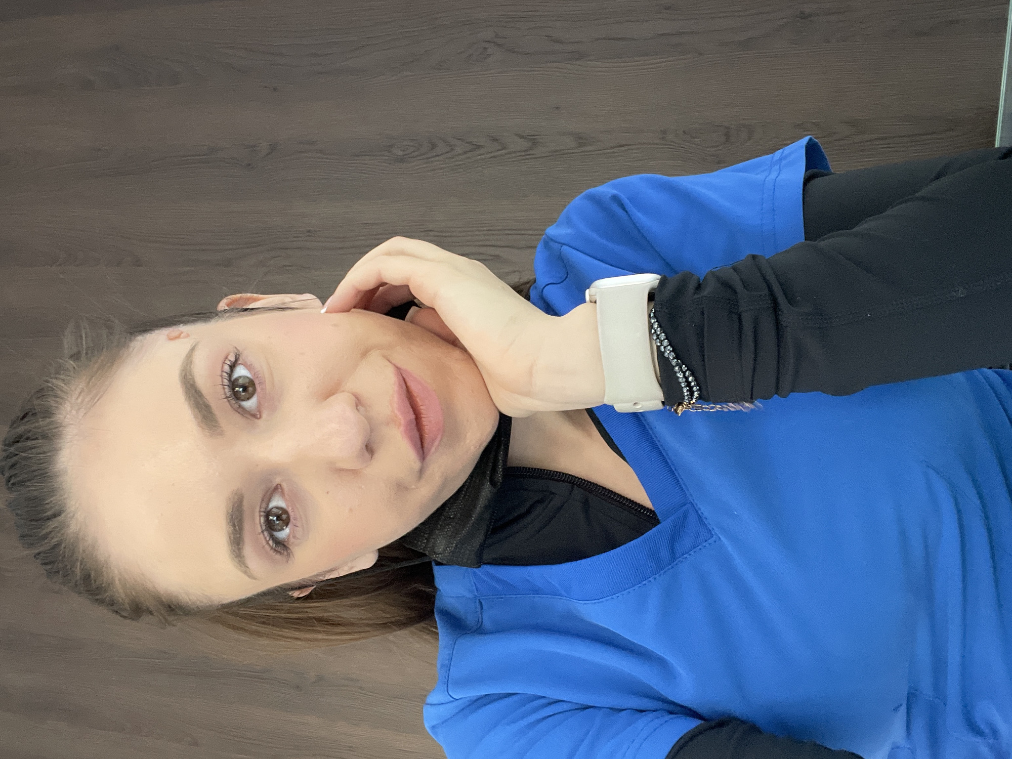 A woman is lying down, facing the camera. She is wearing a bright blue, v-neck top over a black, long-sleeved garment. Her left hand is near her face. She has a light-colored smartwatch on her left wrist and a beaded bracelet. The background appears to be a wooden floor. The location is not evident from the image.