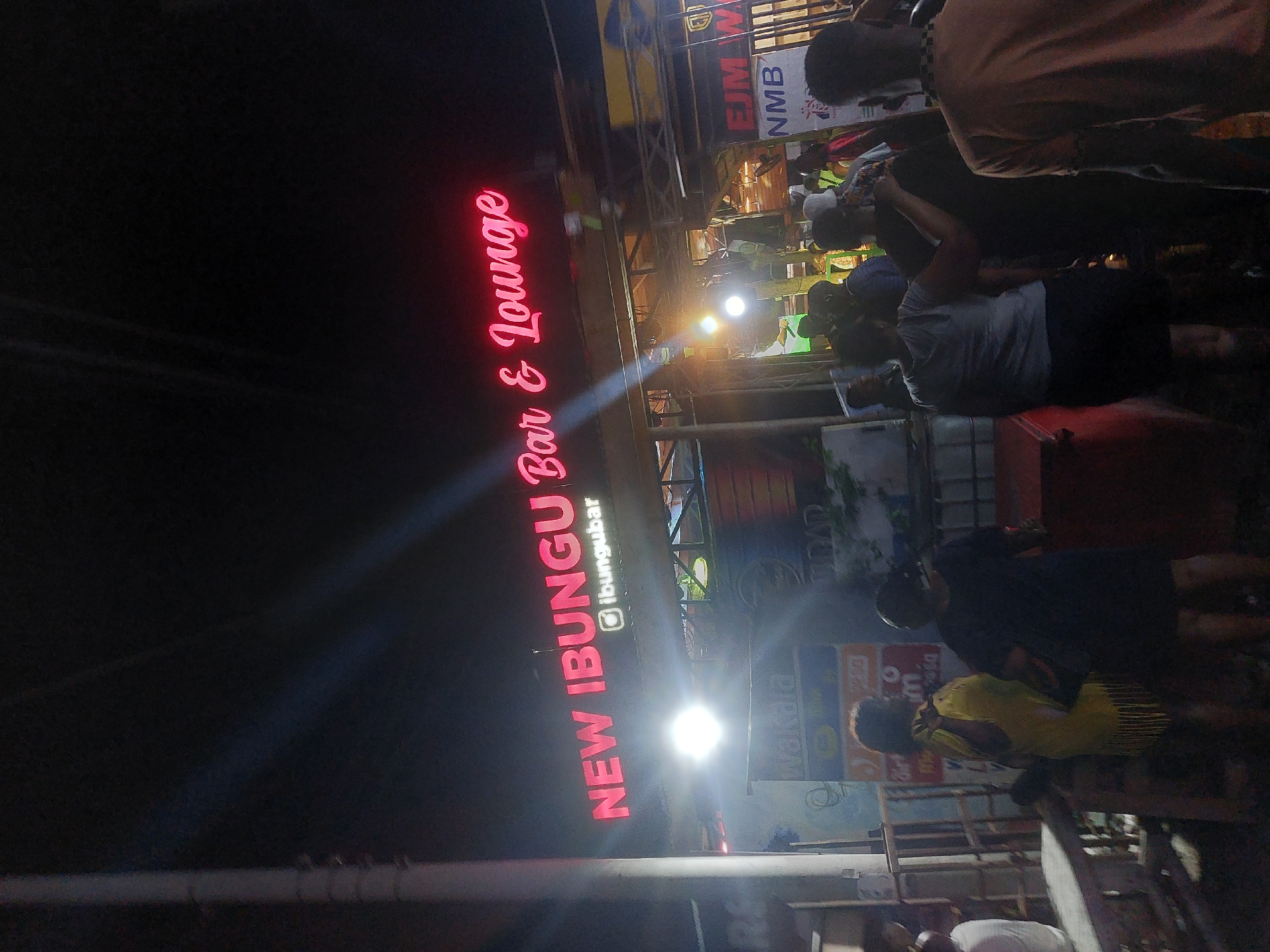The image depicts a nighttime scene outside the "New Ibungu Bar & Lounge." The sign is illuminated in bright red neon lights. Several people are standing and milling about in front of the bar. There are various advertisements and signs visible in the background, and the environment looks to be outdoors. Based on the presence of "NMB" and other local advertisements, the location is likely in Dar es-Salaam, Tanzania.