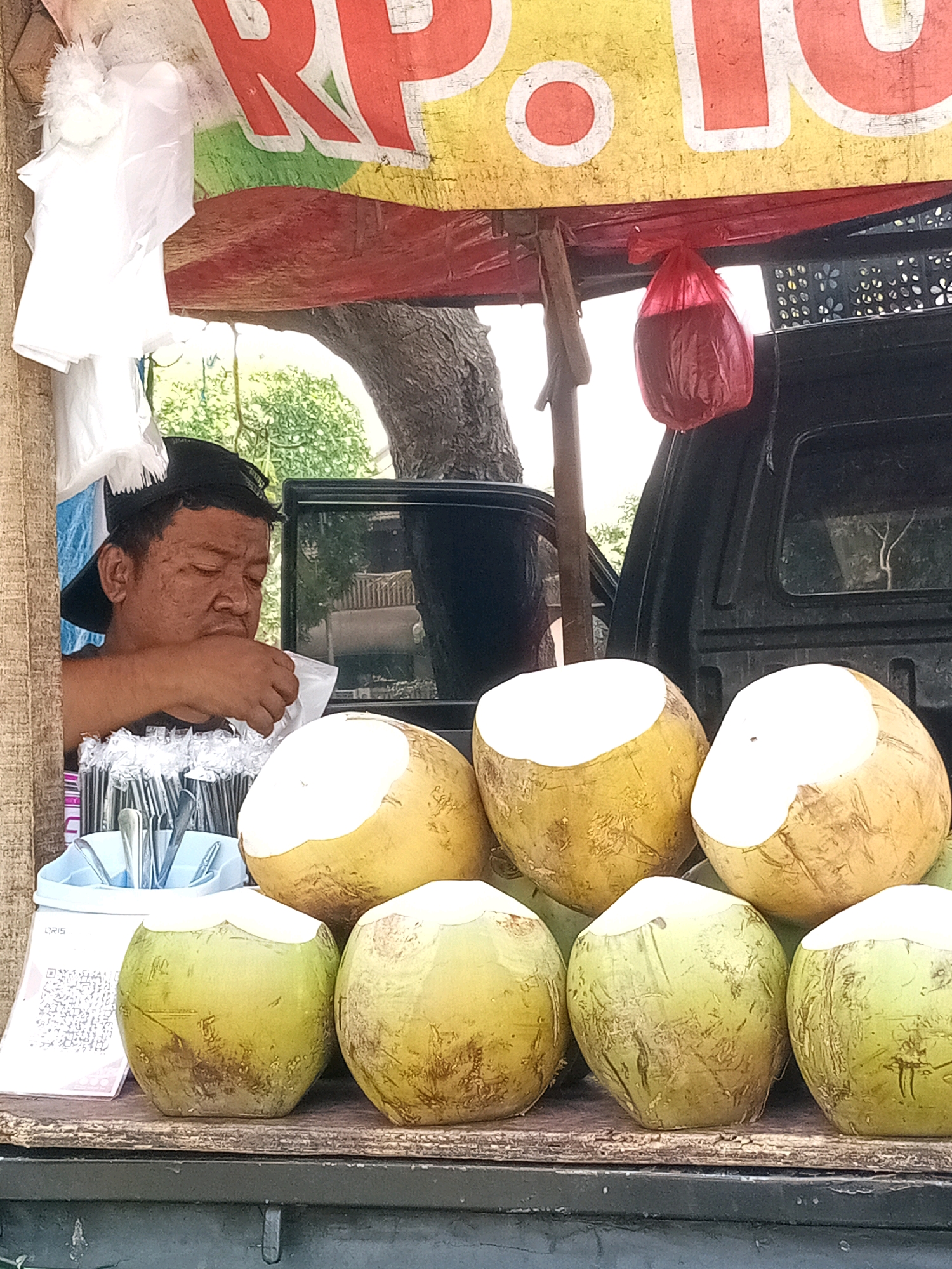 A man wearing a black baseball cap is seen hunched over at a street-side stall, appearing to be preparing or consuming something from a white plastic bag in his hand. He is positioned to the left of a display of fresh coconuts, some of which have their tops cut off, revealing the white flesh inside. The stall features a yellow tarp overhead with red and green lettering that is partially visible. A red plastic bag hangs from the tarp. Behind the man and the coconuts, a dark-colored vehicle, possibly a truck, is parked. The environment appears to be outdoors, with trees and foliage visible in the background. A QR code is visible on a white surface to the left of the coconuts. The scene suggests a vendor at a market or roadside stand in Bekasi, Indonesia, selling coconuts.