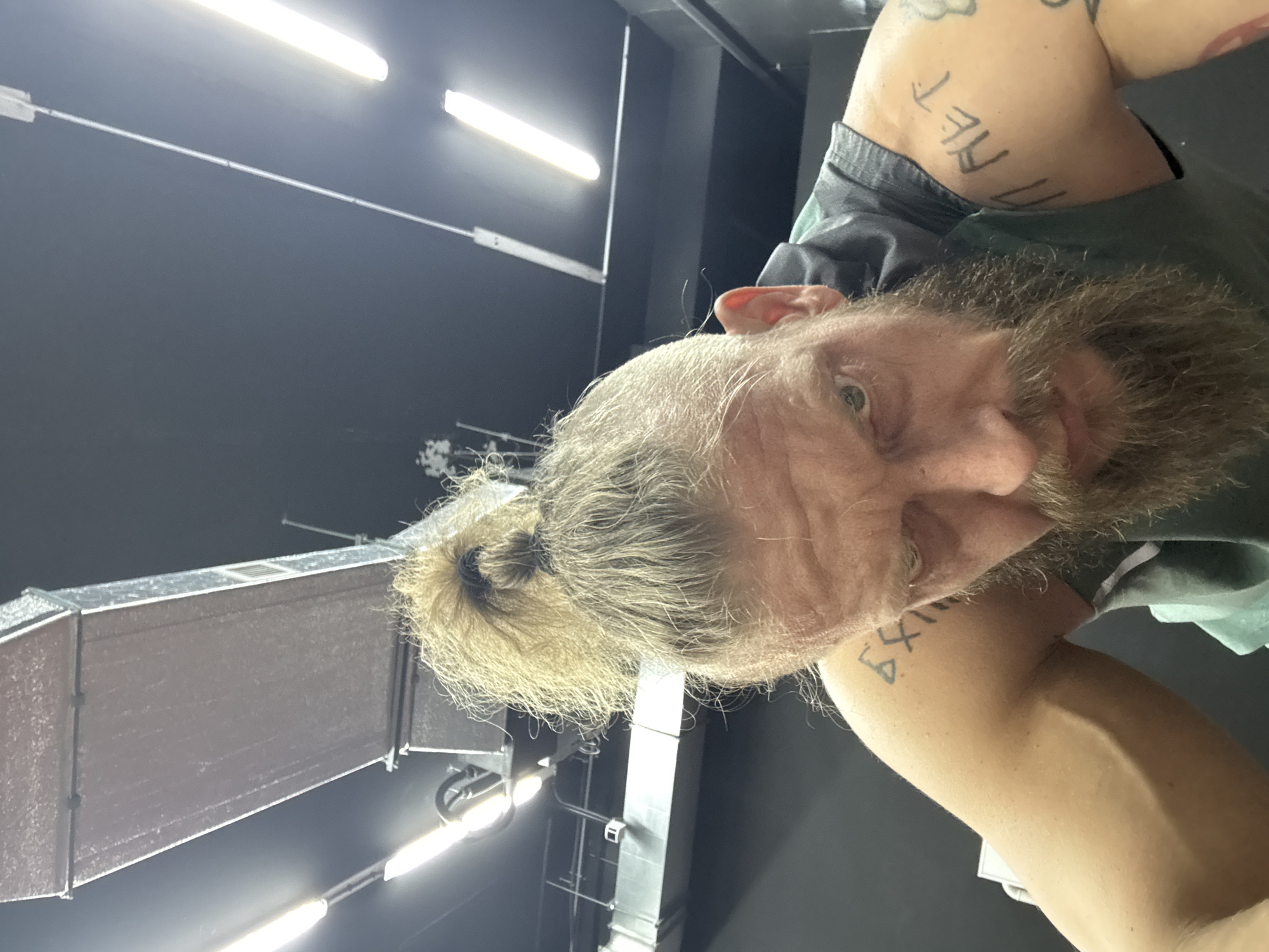 A man with fair skin, a full, light-colored beard, and long light-colored hair tied in a man bun is the primary subject of this close-up, eye-level indoor portrait. He wears a dark t-shirt and looks directly at the camera with a neutral expression. His eyes are blue or green.
Tattoos are visible on both upper arms. On his right upper arm (viewer's left), the words "THE NET" are clearly legible. On his left upper arm (viewer's right), there are several symbols or letters, including what appears to be a "6" and other unidentifiable shapes.
The background reveals a dark ceiling with bright, linear fluorescent tube lights arranged horizontally. Below the lights, a large, rectangular metallic air duct system is visible, extending across the frame. The setting appears to be an indoor industrial-style space, possibly a gym, workshop, or similar commercial building, located in Volzhsky, Russia. The image is originally oriented vertically, rotated 90 degrees counter-clockwise.