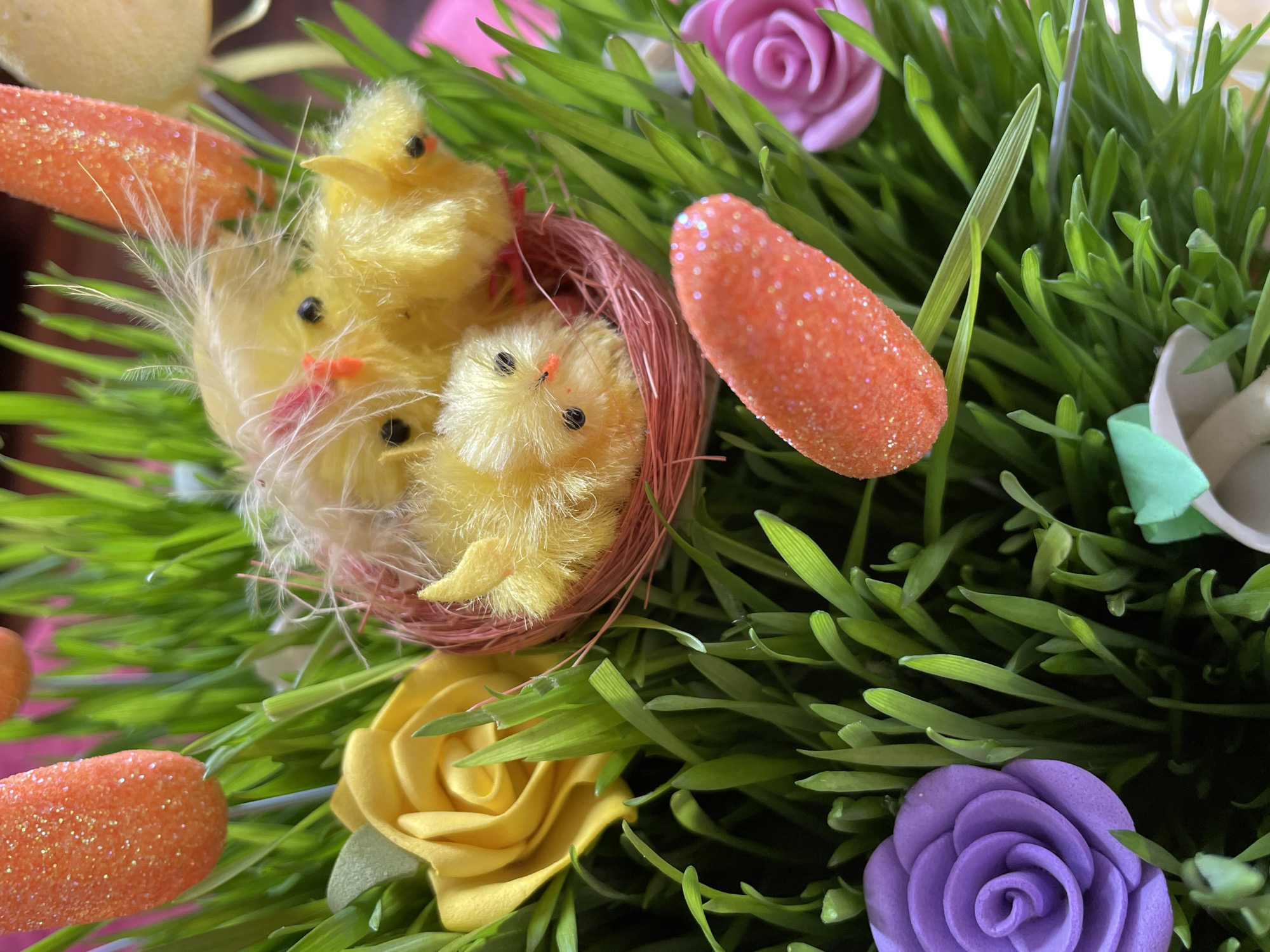 A close-up shot displays a nest filled with fuzzy, yellow chicks amidst lush, green grass. Several orange, glittery carrots and colorful roses are scattered around the nest and grass. The roses are pastel shades of purple, yellow, and pink. The scene evokes a spring or Easter theme. Location information (Aratashen, Armenia) is not visually discernible in the image.