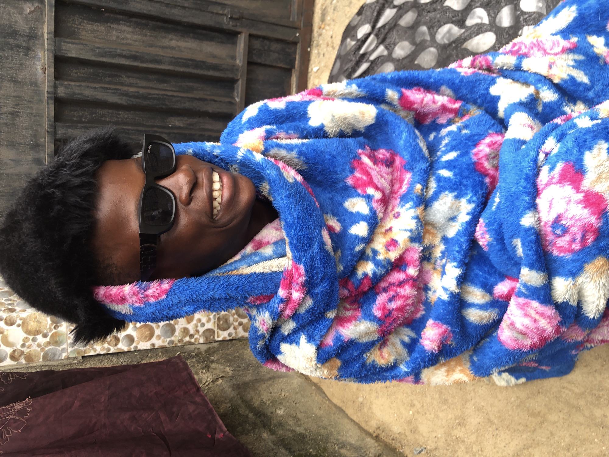 A person wearing a black fur hat and black sunglasses is wrapped in a blue, pink, and white floral blanket. The person is smiling, showing their teeth. They are standing outside near a wall with a patterned surface and a corrugated metal gate. A dark red fabric hangs on the left side of the image, and a black and white spotted cloth lies on the ground to the right. The ground appears to be sandy. The scene is set in Ado, Nigeria.