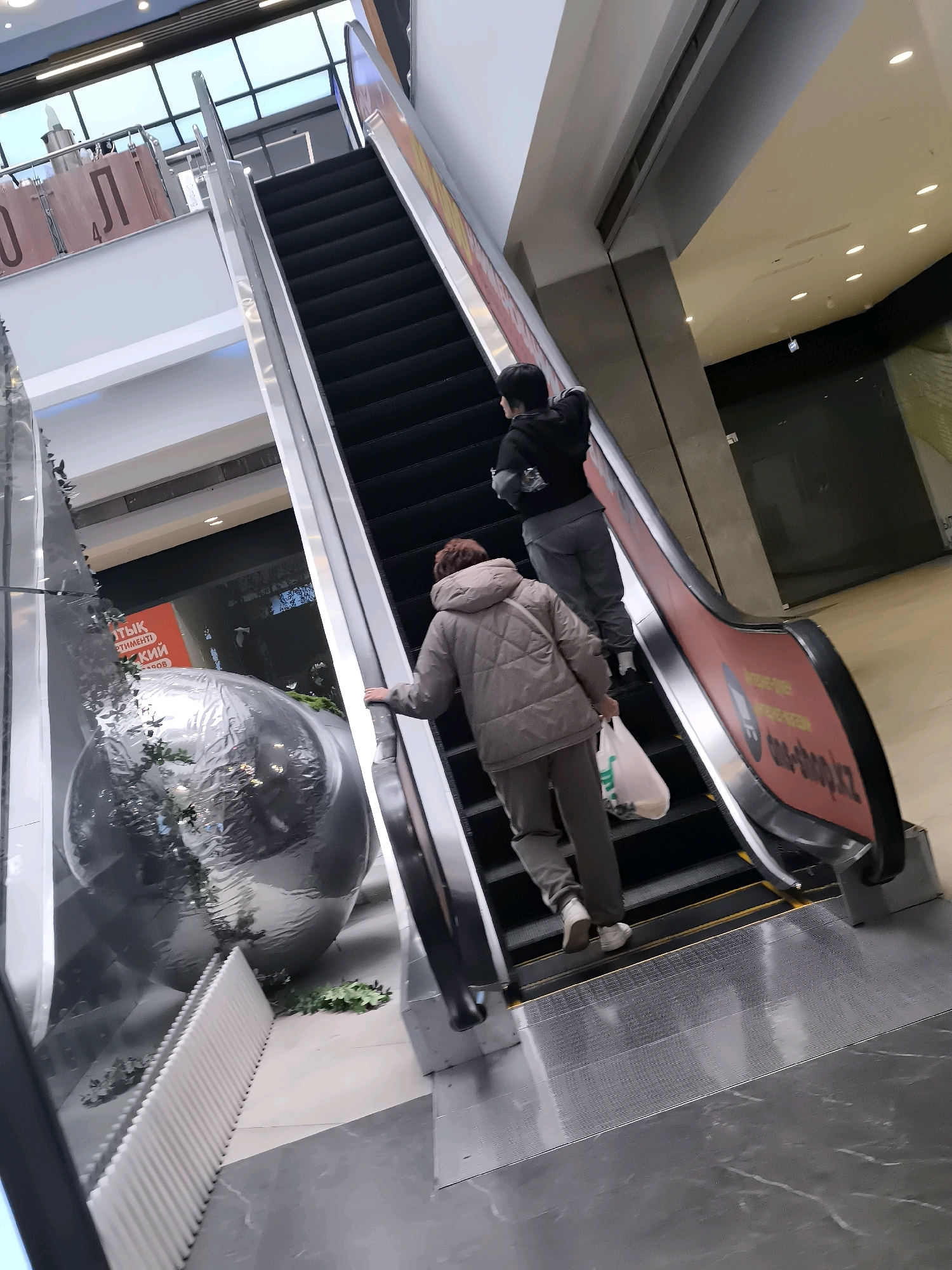 Two individuals are ascending an escalator in an indoor commercial space. The person in front, a woman wearing a quilted grey jacket and grey pants, is carrying a white plastic bag. Behind her, a younger individual, appearing to be a child or teenager, is wearing a black hooded jacket and grey pants, with a dark bag slung over their shoulder. The escalator is dark grey with yellow safety markings. To the left of the escalator, there is a large, reflective, spherical object with green foliage around its base, and further left, a white ribbed structure is visible. The environment appears to be a modern shopping mall or similar establishment. There are indications of signage in Cyrillic script on the escalator and in the background. While the prompt mentions Qarağandı city administration, Kazakhstan, there is no direct visual evidence within the image to confirm this specific location.