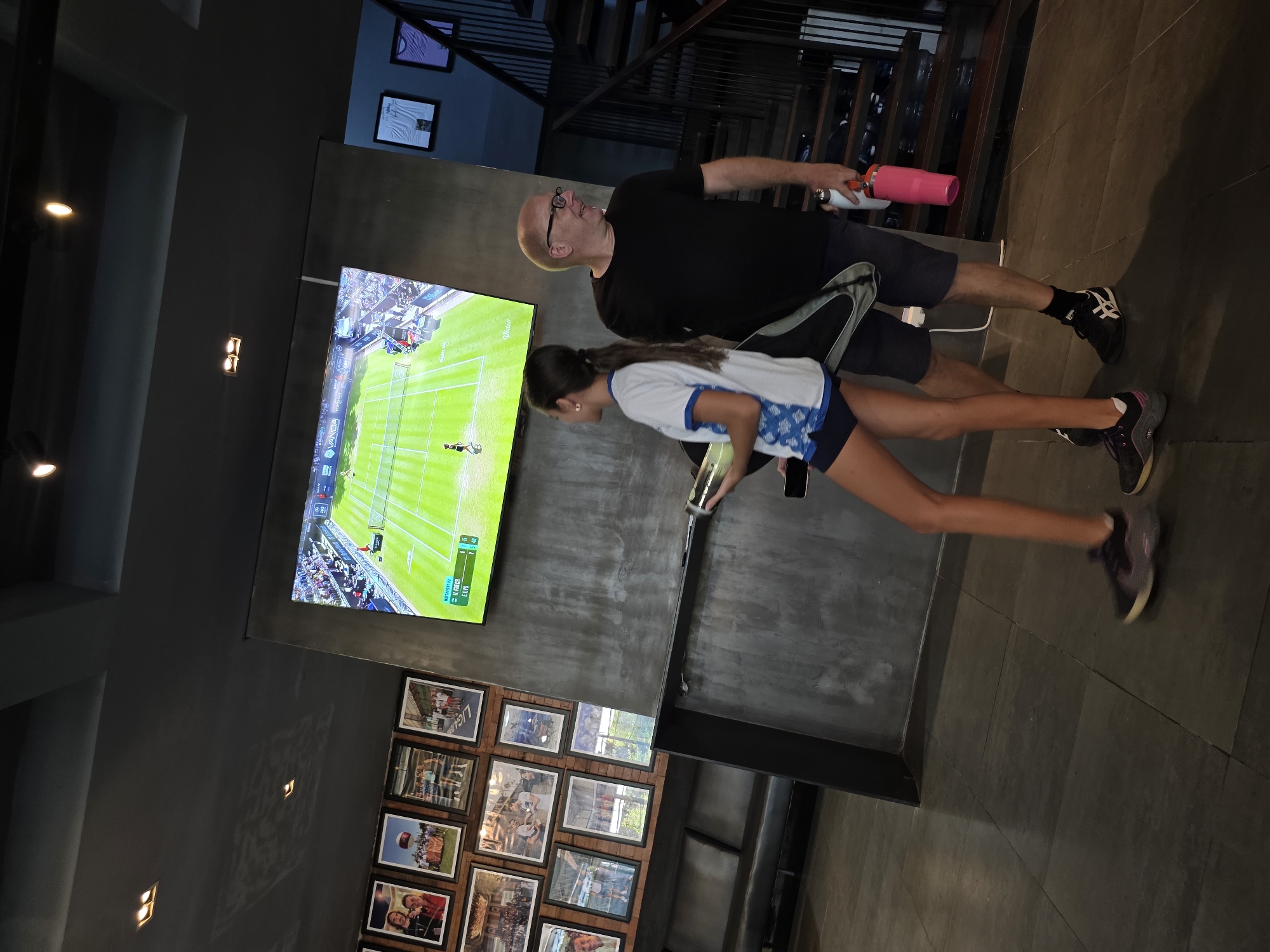 This horizontal media file shows a man and a young woman in what appears to be a modern living space with a grey concrete wall and tiled flooring. They are both standing and facing a large television screen displaying a tennis match. The man, wearing glasses and a black t-shirt, holds a pink water bottle. The woman, in a white and blue top, is looking at a phone she's holding. Framed photographs are mounted on the wall to the left of the television. The scene suggests a casual, indoor activity. The lighting indicates it is likely evening or indoors with artificial light, given the apparent dimness of the surroundings. The presence of framed photos on the wall and the modern decor might suggest a home or a recreational area.