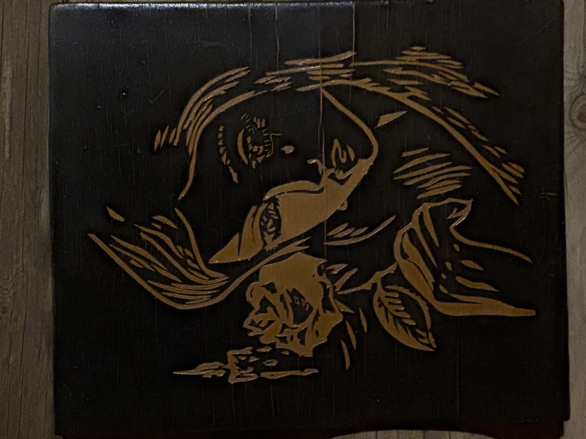 The image shows a dark, likely wooden, surface with a carved or engraved design. The design is in a lighter color, possibly exposing the natural wood beneath the dark surface. The design depicts a stylized woman's head and upper body with flowing hair, appearing to be under an umbrella or canopy. Below the woman, a rose blossom with leaves is included in the composition. The surface appears to be part of a piece of furniture, possibly a stool or box. The background is out of focus, showing a wooden floor.
Маза, Russia, is not discernible from the image.