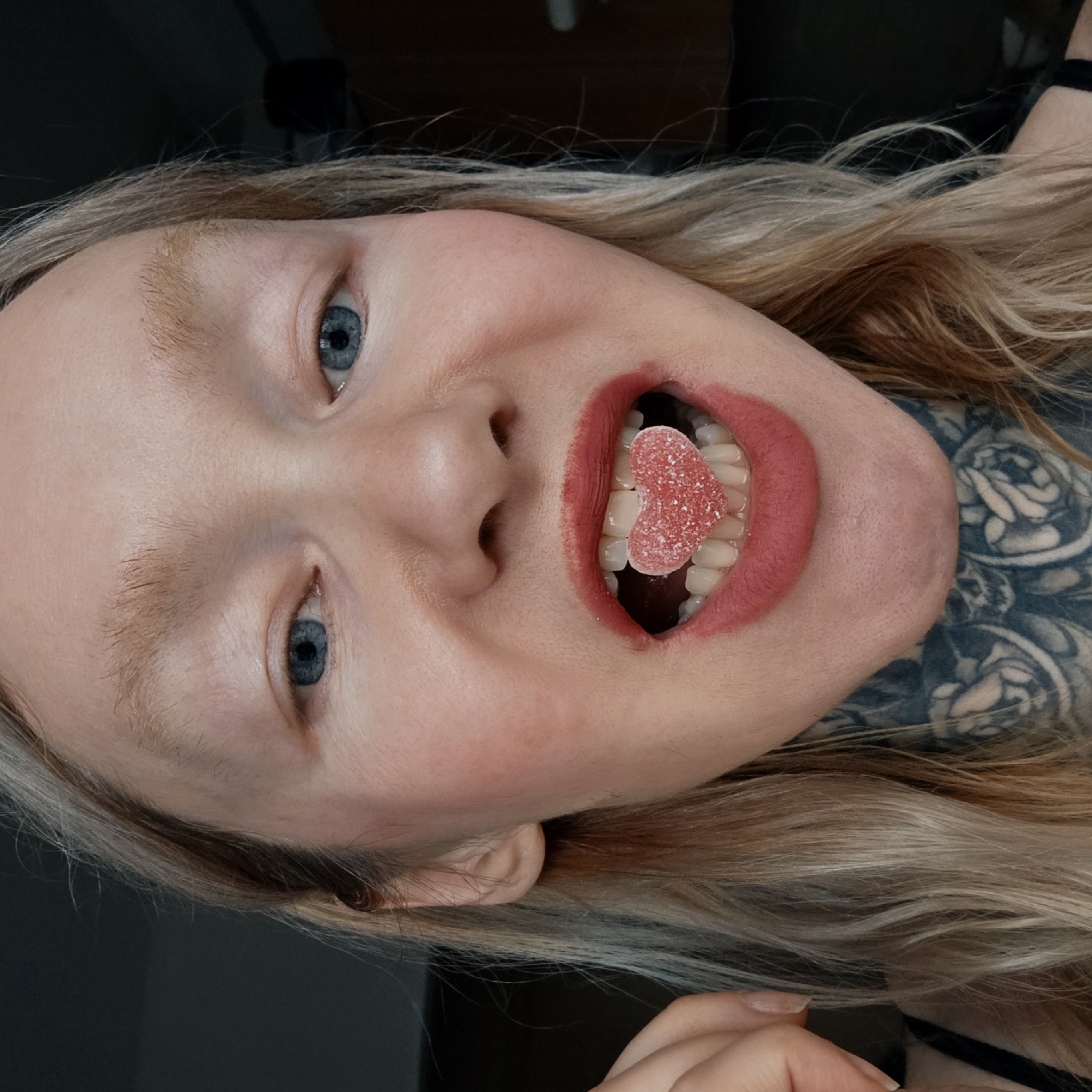 The image displays a close-up, front-facing portrait of an individual with fair skin, light blonde hair, and light blue eyes, looking directly at the viewer. The mouth is open wide, revealing a red, sugar-coated, heart-shaped gummy candy positioned horizontally between the upper and lower central incisors. Red lipstick is applied to the lips, with a distinct vertical smear extending from the center of the lower lip onto the skin below. The individual's eyebrows are light-colored and appear sparse. Extensive black and grey tattoos, featuring intricate floral or skull-like patterns, are visible on the neck and extend down towards the chest. A dark strap, consistent with a garment like a tank top, is partially visible on the lower left. The left hand, with visible fingernails, is present on the far left side of the frame near the ear. The background is dark, out of focus, and undifferentiated, providing no specific environmental details. No identifiable location within the city of Saint Petersburg, Russia, can be determined from the visual information presented.