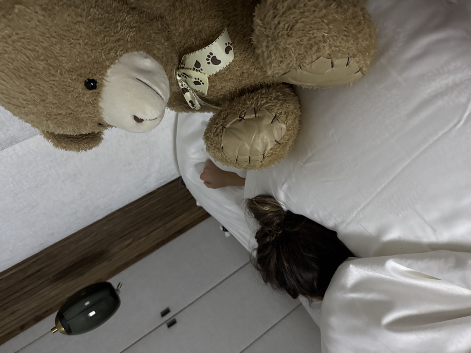 The image depicts an indoor bedroom scene. A large, light brown teddy bear, adorned with a cream-colored ribbon patterned with dark paw prints, is seated upright on white bedding in the right foreground. To the left, a person is partially visible, lying in the bed under white bedding. Only the back of their head, showing dark brown hair styled in an updo, and a portion of one arm are discernible. The person appears to be sleeping. The background features a light-colored paneled headboard or wall behind the bed, and two light-colored closet doors with dark handles to the far left. A dark, tear-drop shaped light fixture with a golden accent is suspended from a wooden beam above the closet area. The specific location within Kastelruth - Castelrotto, Italy, is not identifiable from the visual evidence presented.