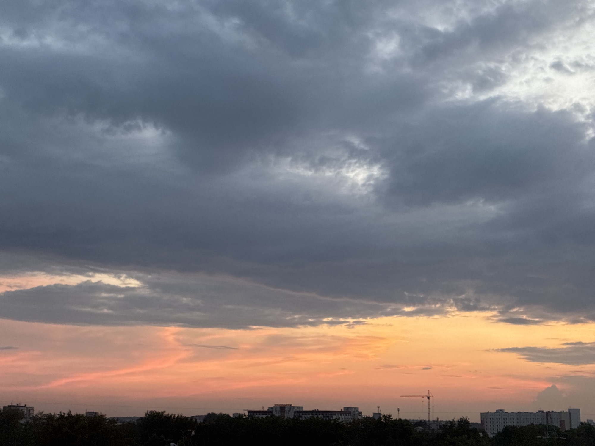 The image depicts a twilight sky over an urban landscape. The sky is filled with dramatic clouds, with dark gray formations dominating the upper portion and transitioning to softer pink and orange hues near the horizon. A strip of dark trees and city buildings runs along the bottom of the frame. Among the buildings, a construction crane is visible on the left side, and several multi-story apartment buildings can be seen. The lighting suggests it is either dusk or dawn.