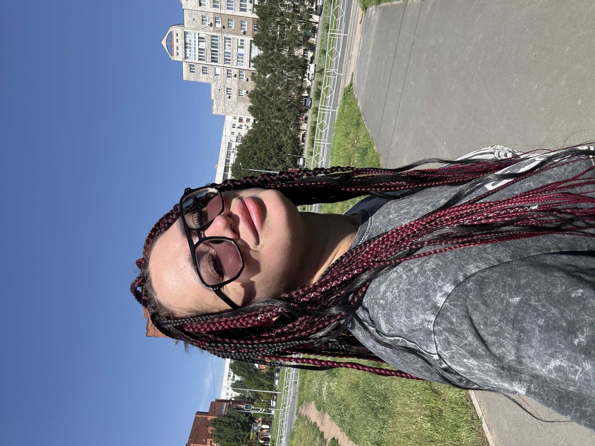 The image is a selfie of a young woman with dark braided hair and glasses, wearing a grey t-shirt. She is standing outdoors on a paved path with grass and trees to her left and a street with cars to her right. In the background, there are several multi-story buildings. The sky is clear and blue. The scene is likely in the city of Tolyatti, Russia.