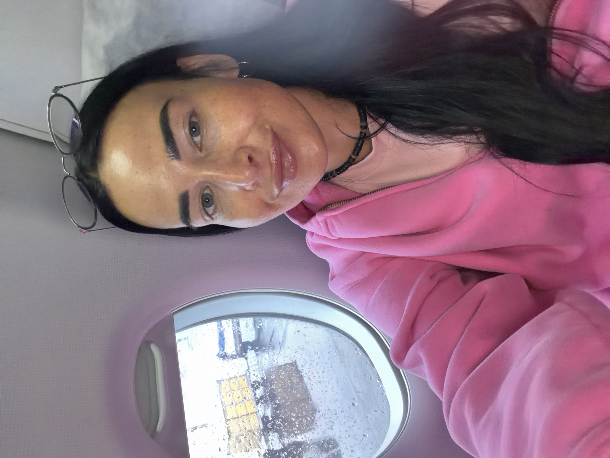 A woman is seated on an airplane, posing for a selfie. She has dark hair, fair skin, and is wearing glasses and a pink hooded sweatshirt. She is wearing a black choker necklace. The window to her left is covered in raindrops, obscuring the view outside. The scene is likely in Khimki, Russia, as the window shows the outside is wet.