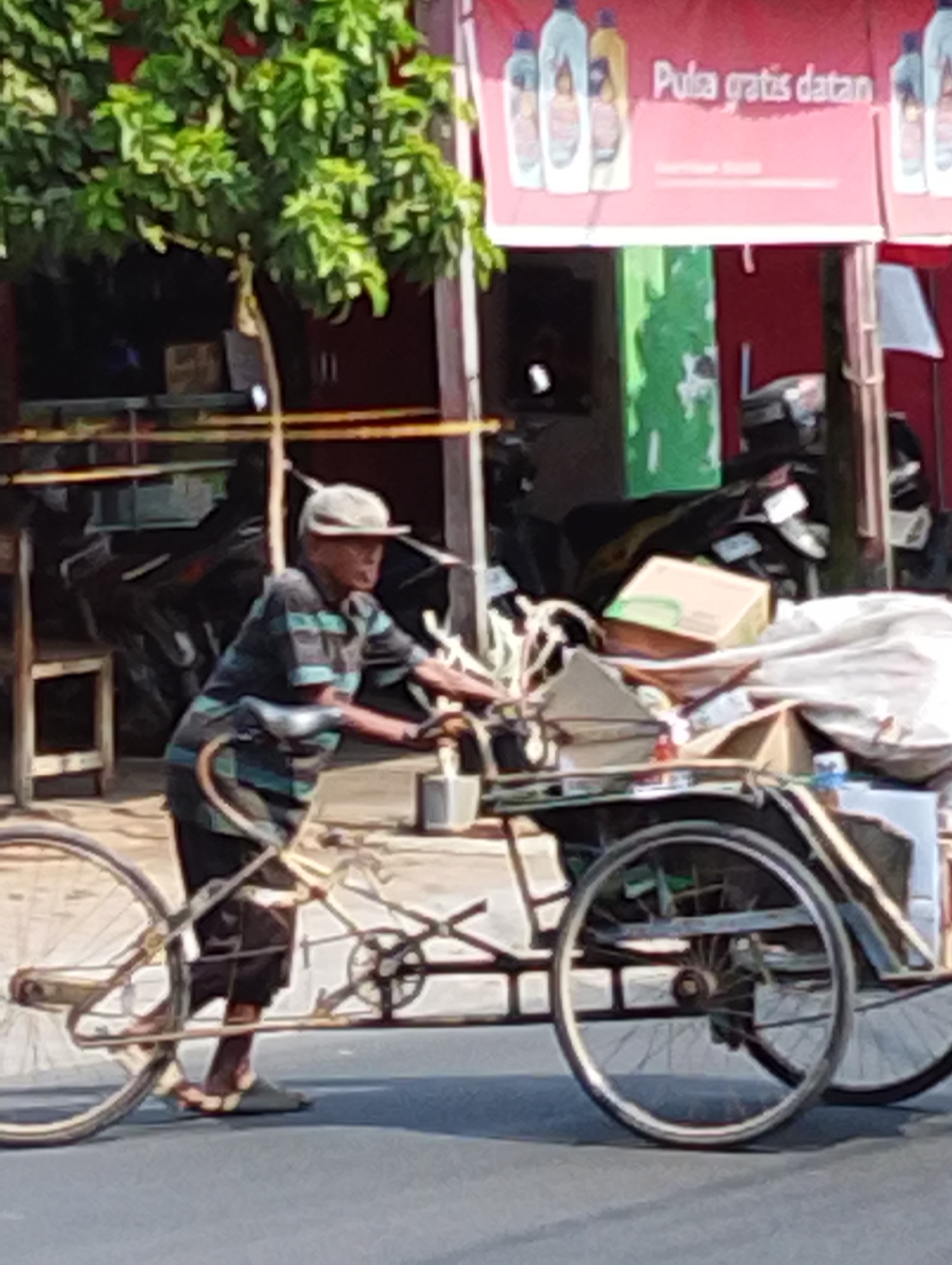 In Manaruwi, Indonesia, a man wearing a striped shirt and a hat rides a bicycle with a cart. The cart is filled with various items, including boxes and what appears to be bundled fabric. In the background, a red banner with the text "Pula gratis datan" is visible, along with a tree and other structures. The scene suggests a street-level view of daily life and commerce in the area.
