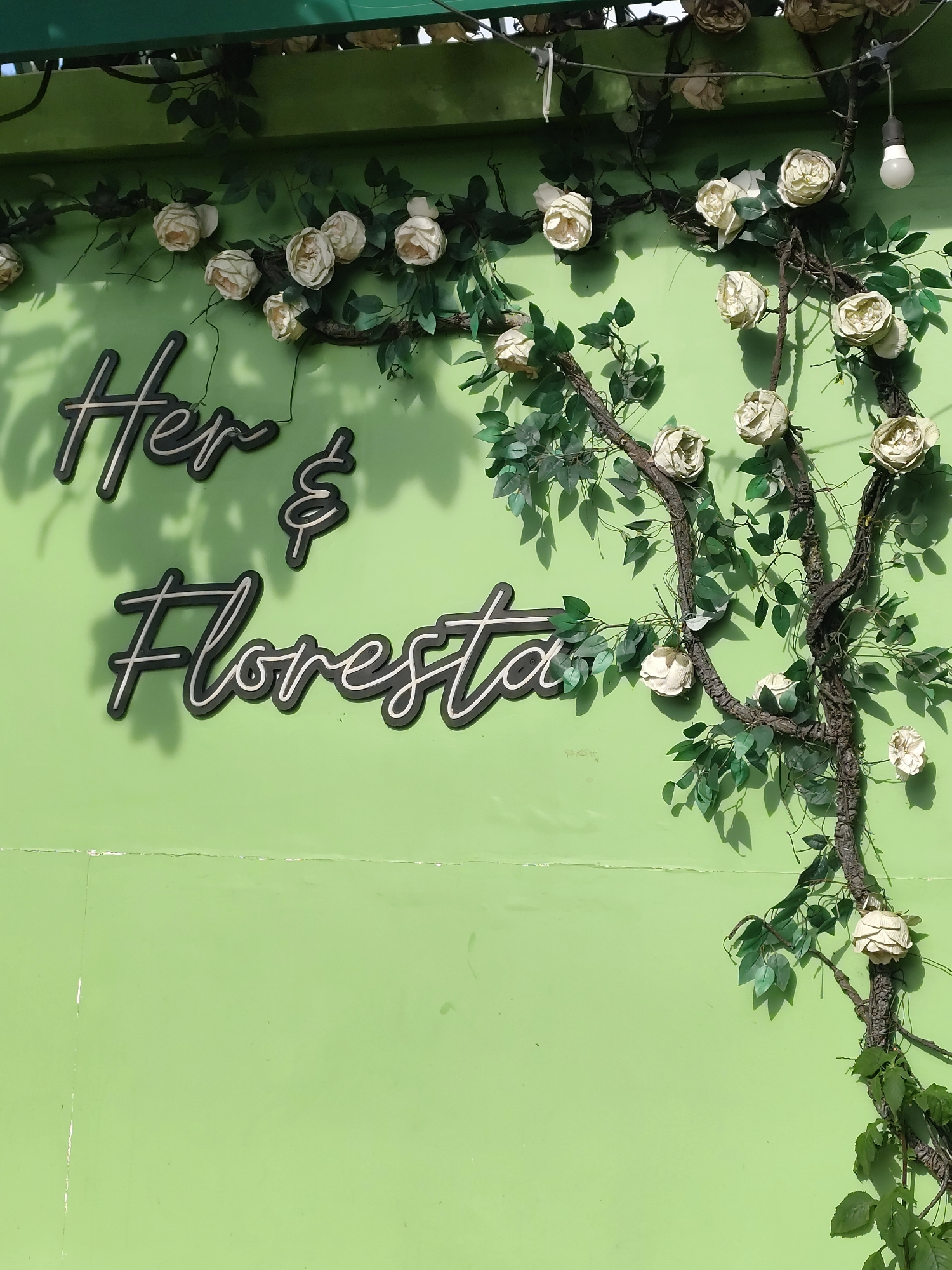 The image shows a light green wall with a neon sign that reads "Her & Floresta." The sign is made of black lettering with a white outline. Artificial green vines with white roses are draped across the wall and the sign. The setting appears to be outdoors, possibly a storefront or cafe in Da Lat, Vietnam, given the signage and floral decoration. There are no people or animals visible in the image. The lighting suggests it is daytime, with shadows from the vines and sign cast on the wall.
