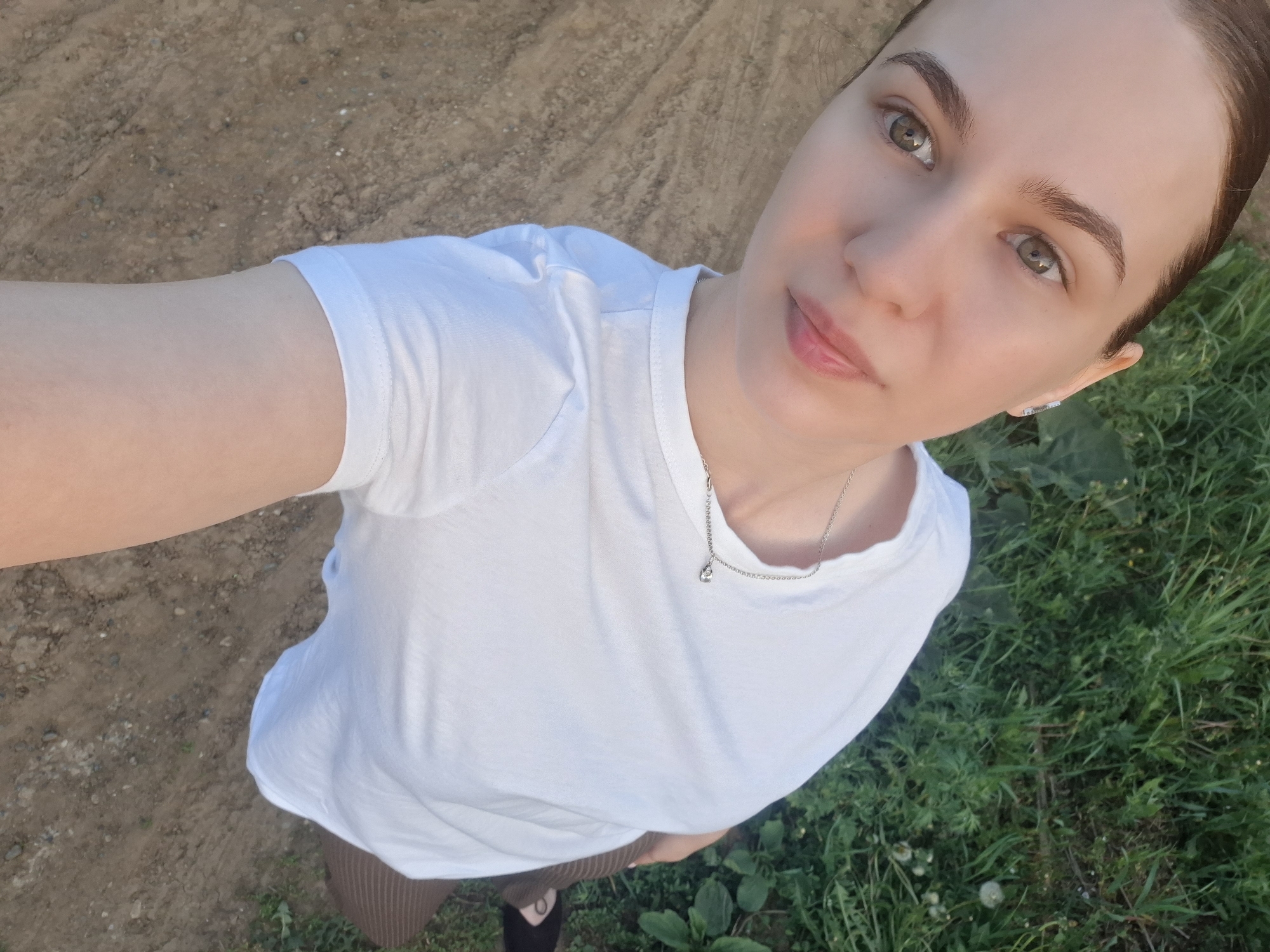 A woman is standing outside in Nizhnyaya Gar', Russia. She is wearing a white T-shirt and a silver necklace. She is wearing brown pants. The ground is a mix of dirt and grass.