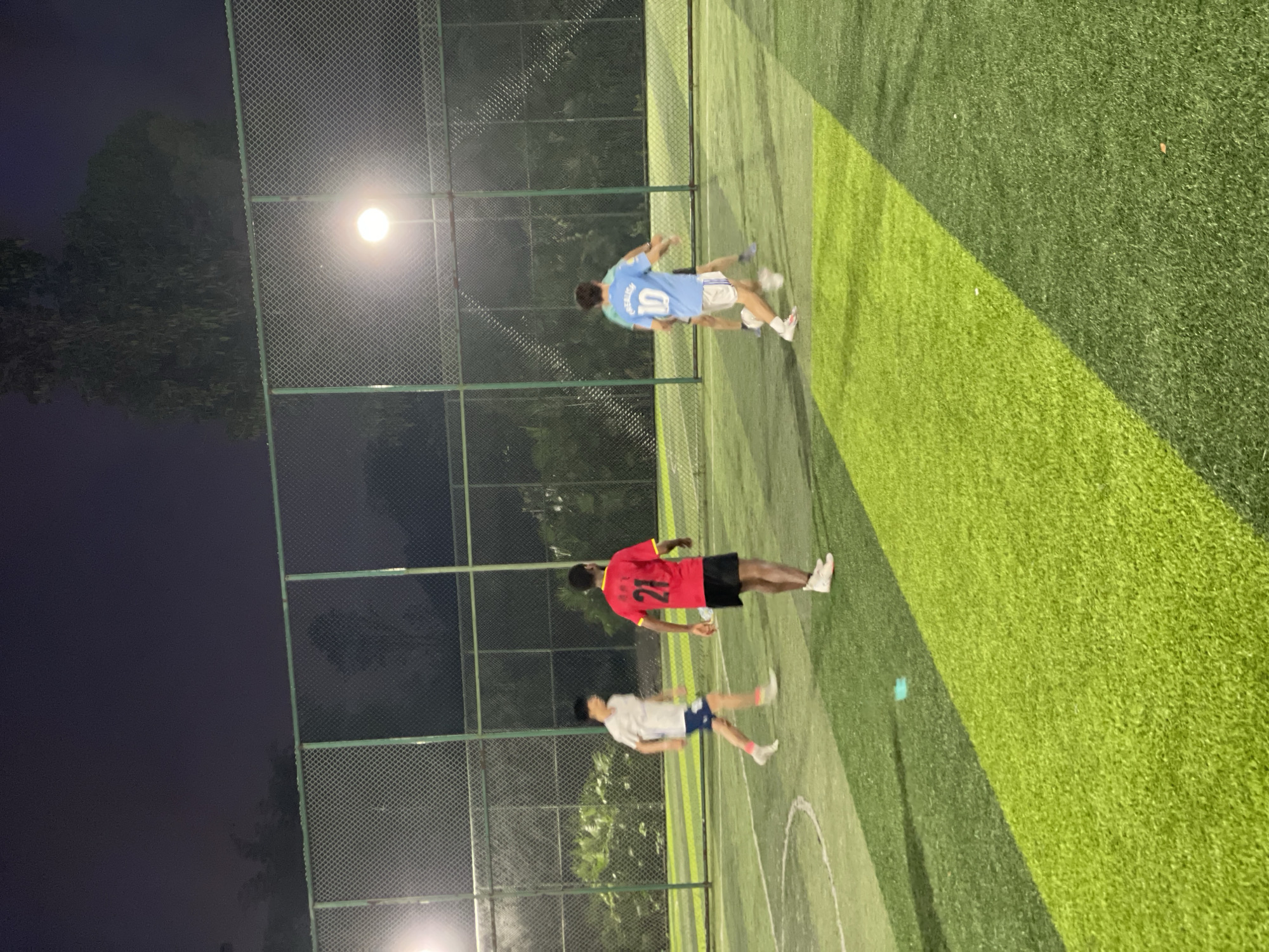 The photograph shows three individuals playing on a soccer field in Jiangjin, China. The scene is illuminated by artificial lights, suggesting it is nighttime. A chain-link fence surrounds the field.
One individual is wearing a light blue jersey with the number 10 on the back and white shorts. Another player is wearing a red jersey with the number 21 on the back and black shorts. The third person is wearing a white jersey and navy blue shorts. The people are in motion. The field has different shades of green grass.
