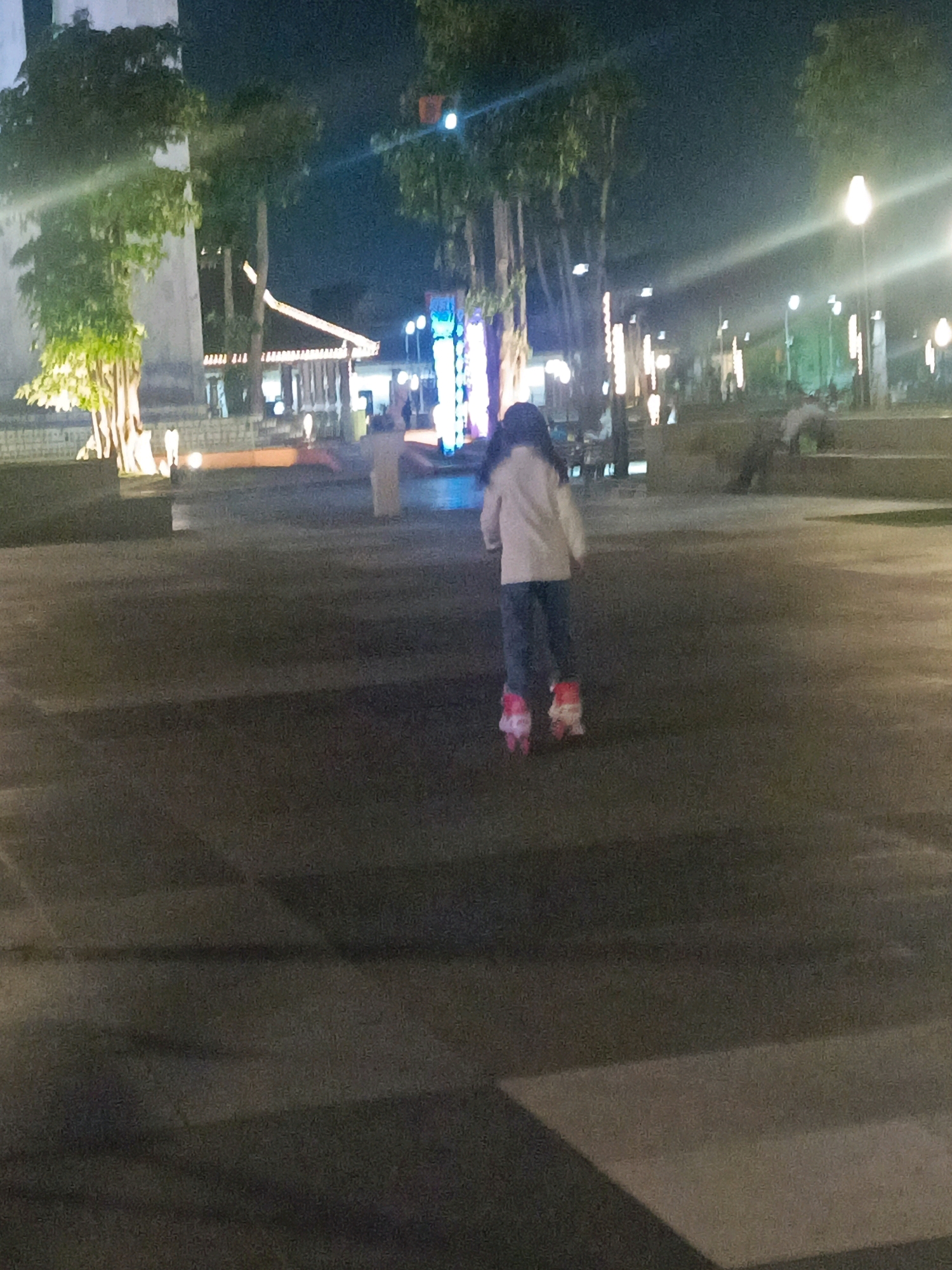 A child is roller skating at night in what appears to be a public park or promenade in Sidoarjo, Indonesia. The child, wearing a light-colored jacket and dark pants, is the central subject and is facing away from the camera. They are wearing pink roller skates. The environment is illuminated by streetlights and decorative lights, casting a warm glow on the scene. Trees line the background, and there are structures that suggest a public gathering space, possibly with a building or pavilion in the distance. Other individuals are visible in the background, though they are less distinct due to the low light conditions and the focus on the child. The ground is paved, and there are some elevated areas or steps in the background. The overall impression is a casual evening outing in a well-lit urban outdoor space.