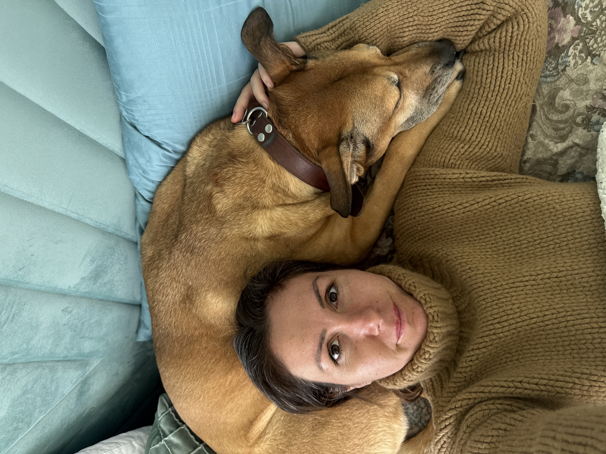 A woman lies on her back, possibly on a couch or bed, with a medium-sized brown dog nestled closely beside her. The dog is positioned so its head rests near the woman's shoulder, and she appears to be cradling the dog’s head. The woman looks up toward the camera, and she is wearing a brown turtleneck sweater. The dog has a brown leather collar with metal hardware. Behind them are cushions, one of which is a light blue color. The specific location within the city of Byala Slatina, Bulgaria, is not discernable.