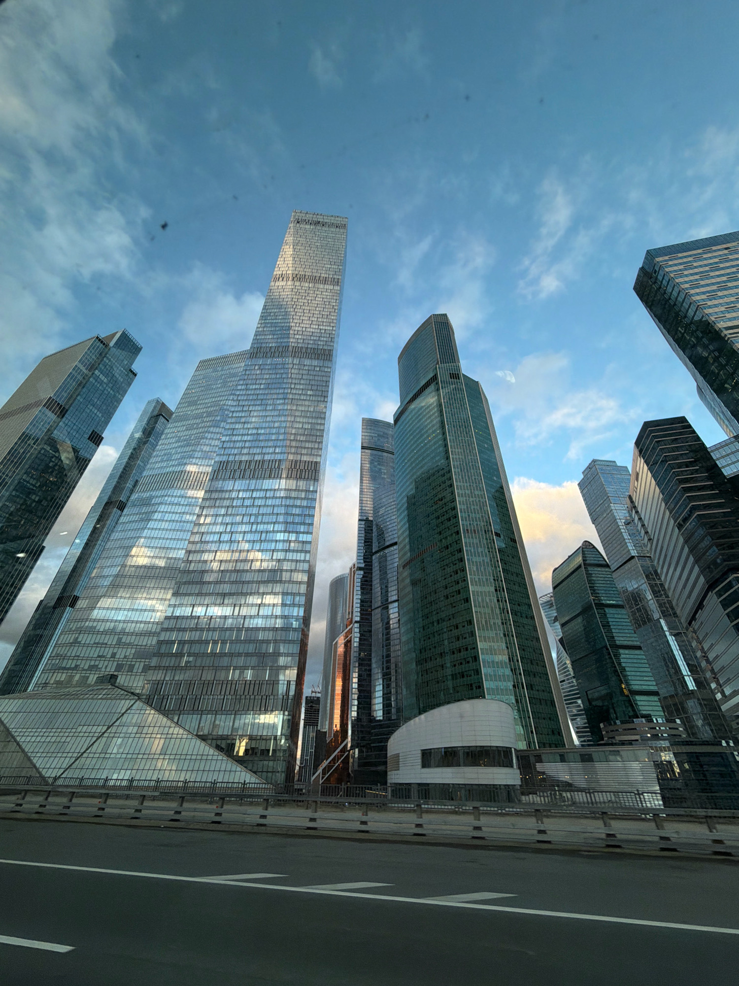 This is a low-angle, wide-shot photograph of the Moscow International Business Center, commonly known as Moscow City, in Moscow, Russia. The image captures a cluster of modern skyscrapers with glass facades, reflecting the blue sky and scattered white clouds. The buildings are of varying heights and architectural styles, with some featuring sharp angles and others a more rounded form. In the foreground, a section of a road or bridge is visible, with a concrete barrier and metal railings separating it from the surrounding structures. The lighting suggests it is either early morning or late afternoon, with sunlight casting warm hues on some of the building surfaces. There are no people visible in the image. The overall impression is one of urban grandeur and contemporary architecture.
