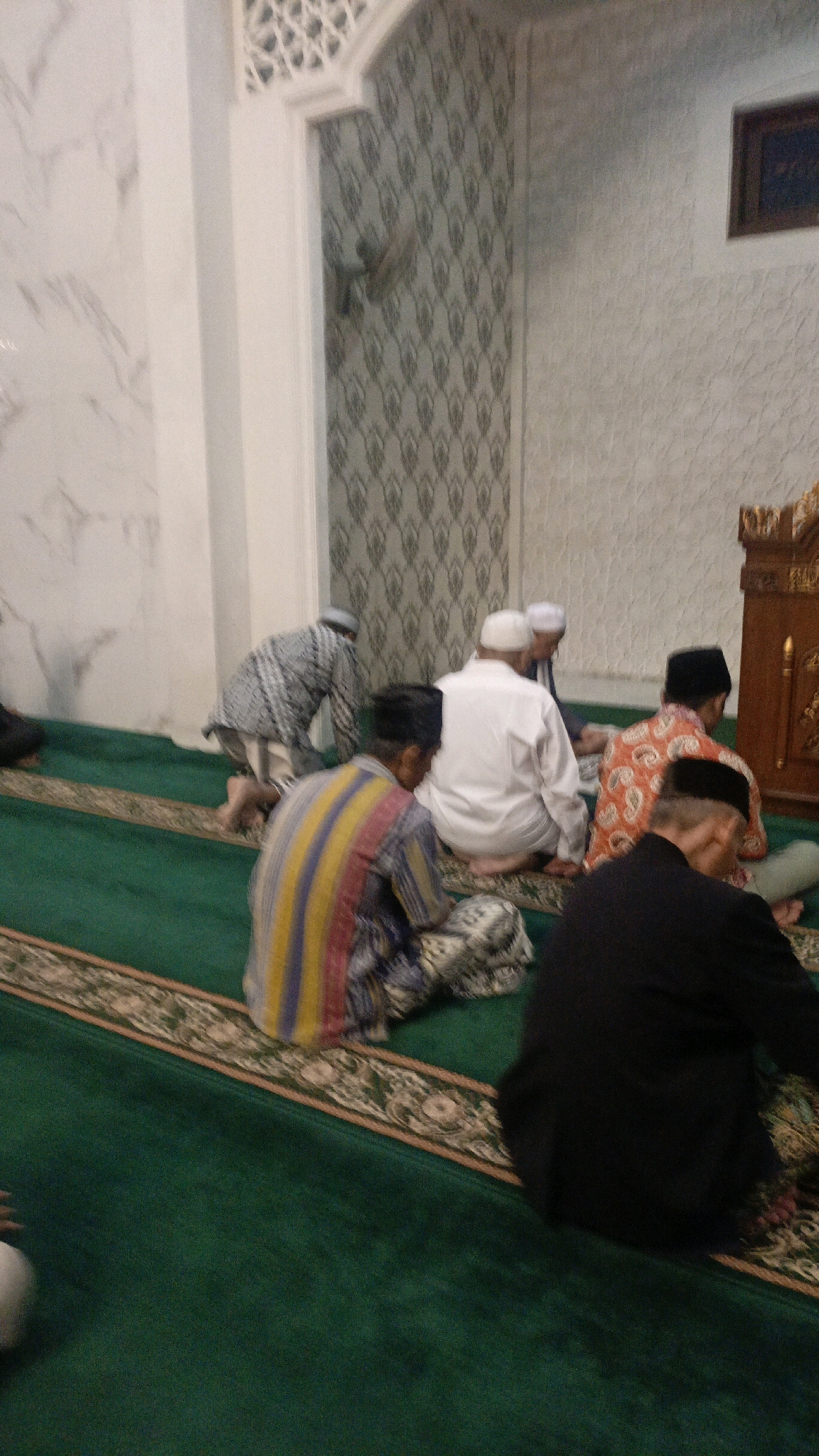 The image depicts a group of men praying inside a mosque. They are seated on a green carpet with patterned borders. The men are dressed in traditional Islamic attire. One man in the foreground is wearing a black jacket and a black cap. To his left, another man is wearing a striped shirt and a patterned cloth. Further back, several other men are visible, some wearing white robes and skullcaps, and one wearing an orange and white patterned shirt. The mosque interior features patterned wallpaper and decorative architectural elements, including a wooden pulpit. The overall scene suggests a religious gathering or prayer service.