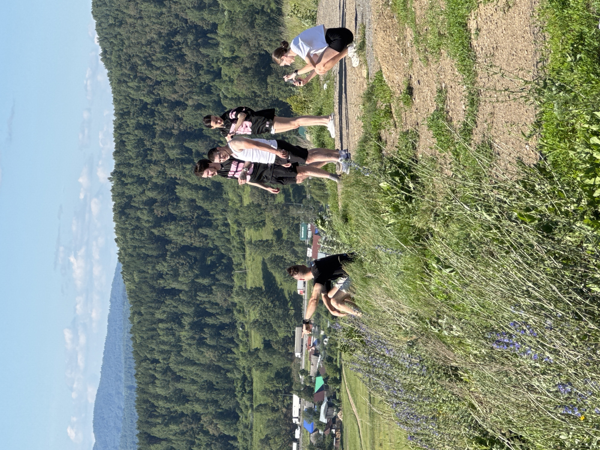 This image captures a scenic overlook in the city of Sim, Russia, on a bright, sunny day. In the foreground, lush green grass and wild purple flowers grow on a gentle slope. A man sits on the grass, pointing towards the valley below, which features a village with houses and a road with trucks. Behind the village, a dense forest covers the rolling hills, with a clear blue sky overhead. On the dirt path to the right, four young adults are gathered. Two men and a woman stand together, while another woman crouches down, taking a photo with her phone. The scene evokes a sense of outdoor recreation and appreciation for the natural beauty of the region.