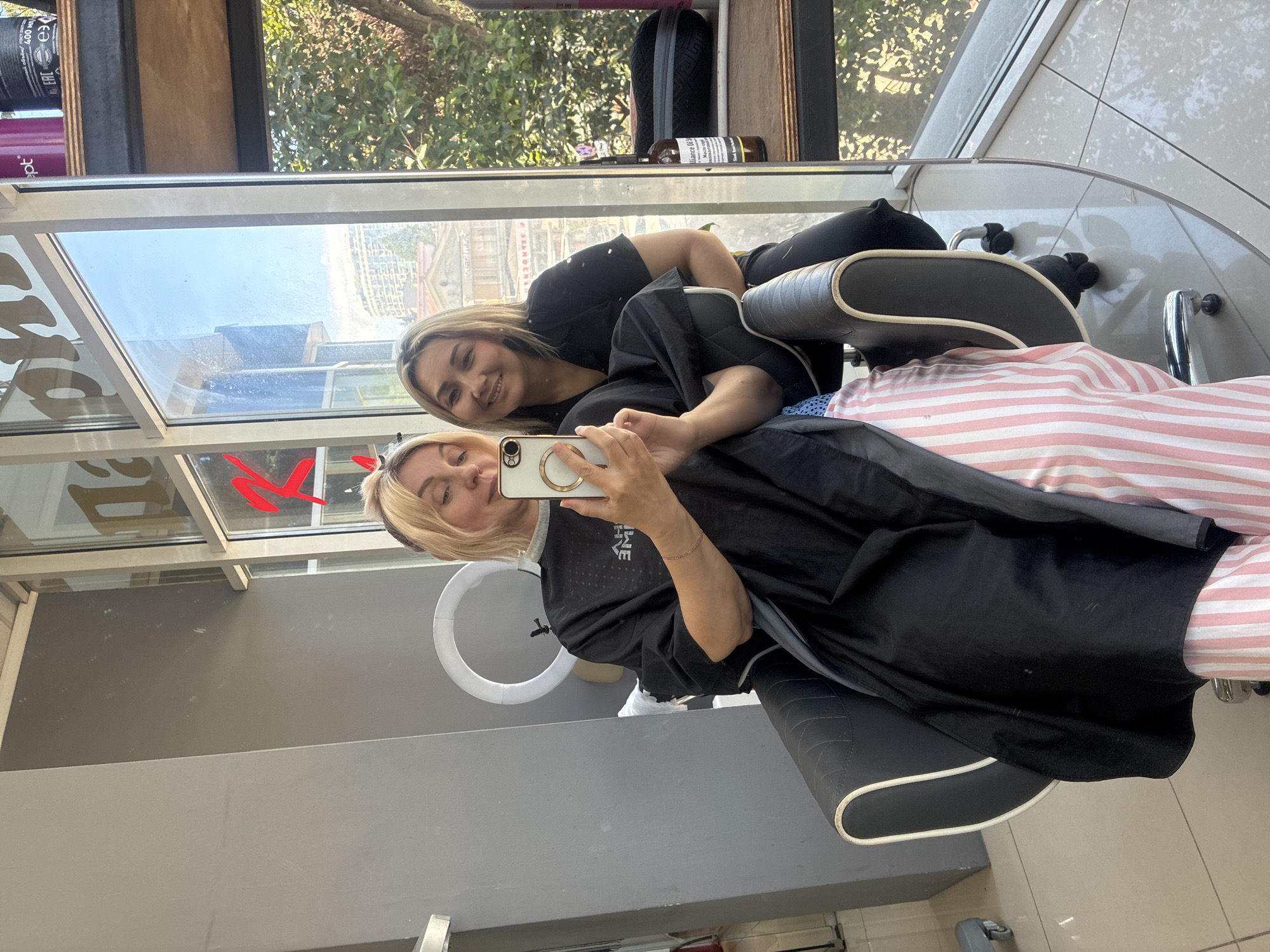 This portrait-oriented image features two smiling women taking a selfie inside what appears to be a hair salon in Sochi, Russia.
In the foreground, a blonde woman, likely a client, is seated in a black salon chair. She is covered by a black cape over a pink and white striped covering. She holds a white smartphone with a gold ring stand in her right hand, taking the selfie, and displays a subtle smile. A small red decorative element, possibly a bow, is visible in her hair near her left temple. Standing closely behind her and leaning into the frame is a second woman, presumably her stylist. She has blonde-highlighted hair, a warm, broad smile, and looks directly at the camera. She is wearing a black top.
The background is dominated by a large window that offers a view of a sunny outdoor scene. Through the window, lush green trees, a clear blue sky, and a light-colored, possibly old, building with distinct architectural details are visible. The window also reflects interior elements of the salon, including a prominent white circular ring light positioned near the client's head, and a faint red stylized letter or logo.
On a shelf visible through the window, several bottles are present. One purple bottle shows part of the word "Opt," while a black bottle is marked "400 мл" and features an "EAC" logo. Another brown bottle has partially legible text, possibly "Brilliance of Sl..." or "Balance of Sl...".
The bright natural light suggests it is daytime, likely a clear and sunny day. Both women appear cheerful and happy, conveying a pleasant and friendly atmosphere within the salon.