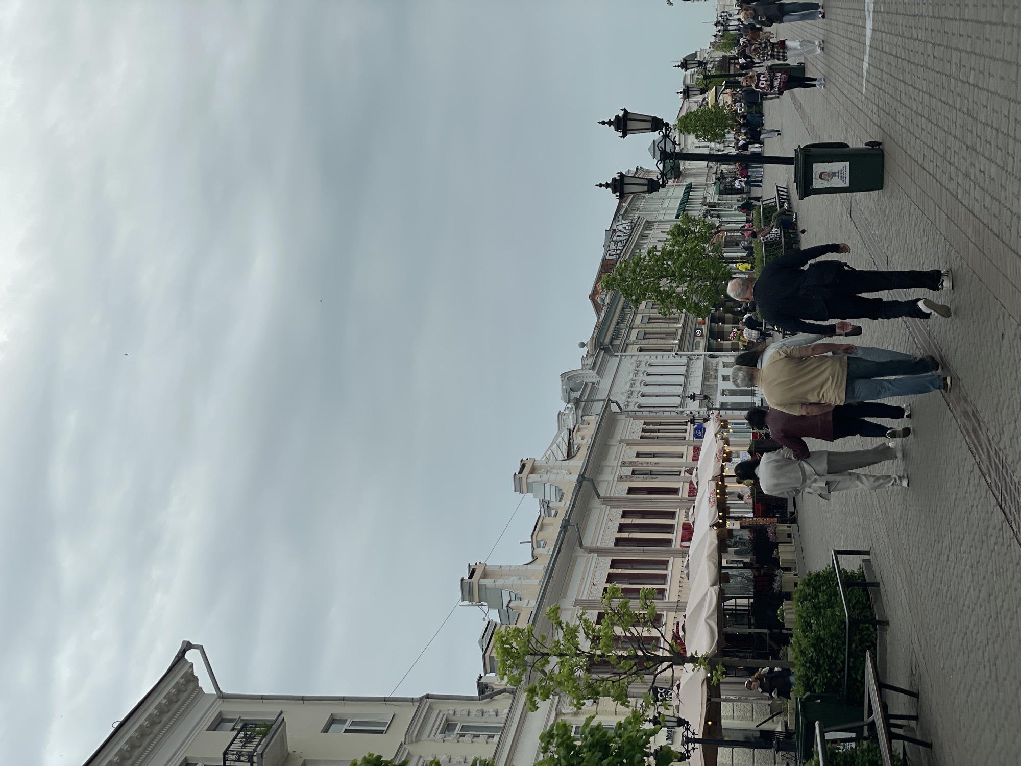 The image shows a pedestrian street in Kazan, Russia, under a cloudy sky. The street is lined with buildings, many featuring ornate architectural details. Several pedestrians are walking along the paved sidewalk, with some seated at outdoor cafes with white umbrellas. There are lamp posts on the street. A green trash bin is also visible on the sidewalk.